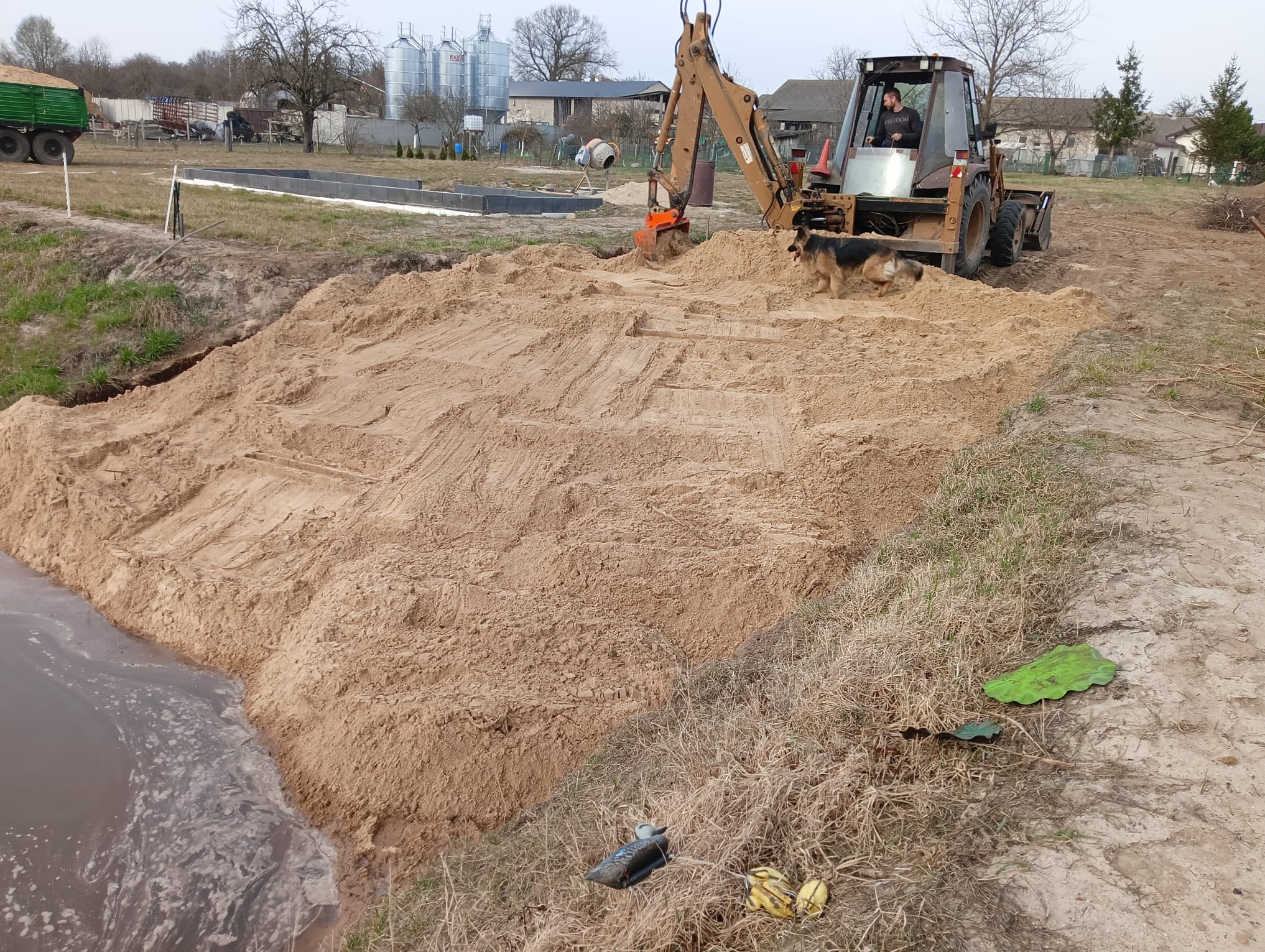 Żółta koparko-ładowarka na piaszczystym terenie, obok zbiornik wodny. W tle budynki gospodarcze i silosy. Pies rasy owczarek niemiecki przy maszynie.