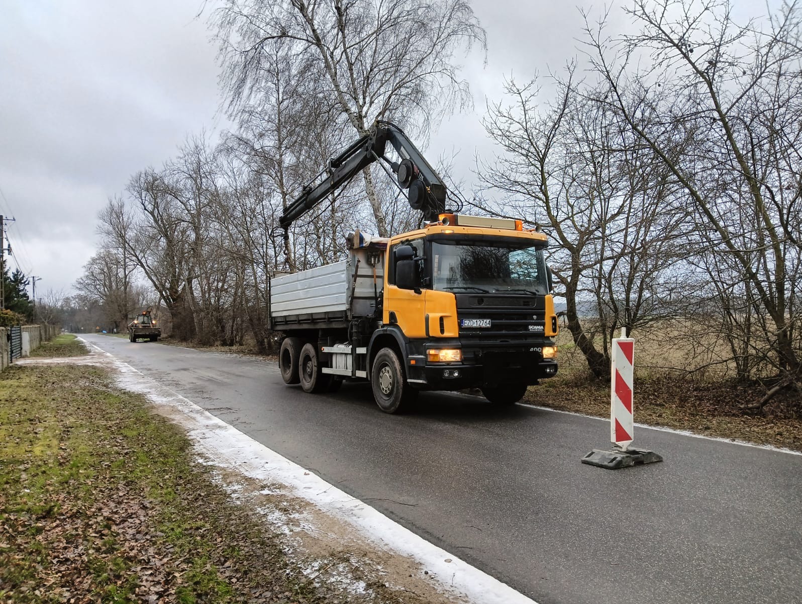 Żółta ciężarówka z dźwigiem w Szadku na drodze, w tle drzewa bez liści i inny pojazd. Widoczny znak drogowy.