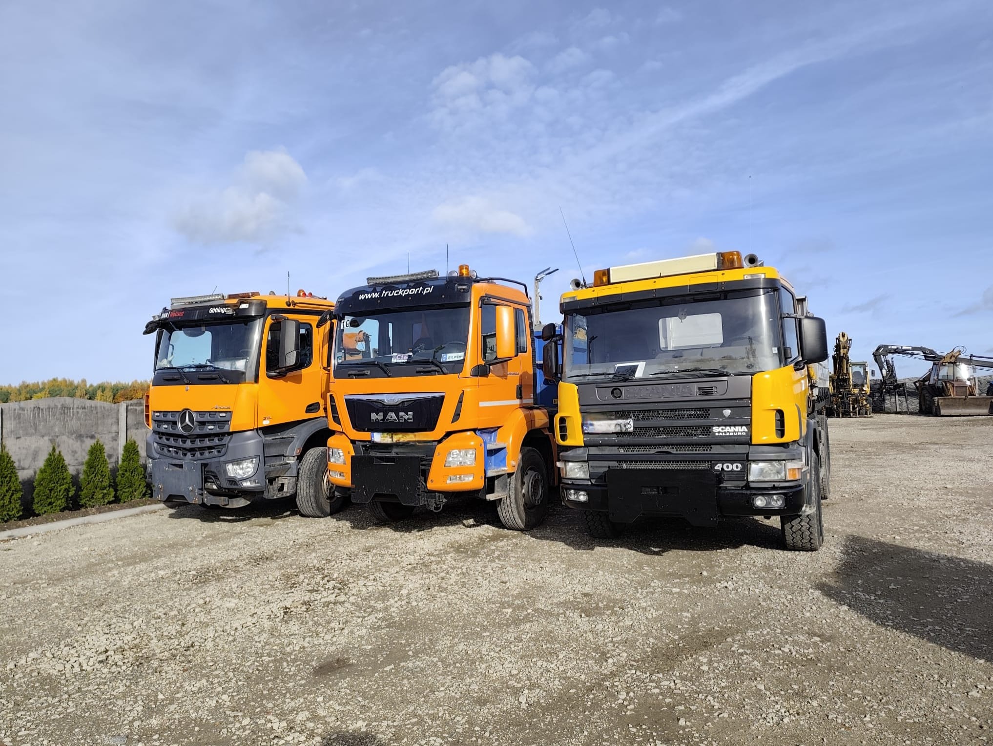 Trzy pomarańczowo-żółte ciężarówki (Mercedes, MAN, Scania) stojące na żwirowym placu, z koparkami w tle. Błękitne niebo z chmurami.