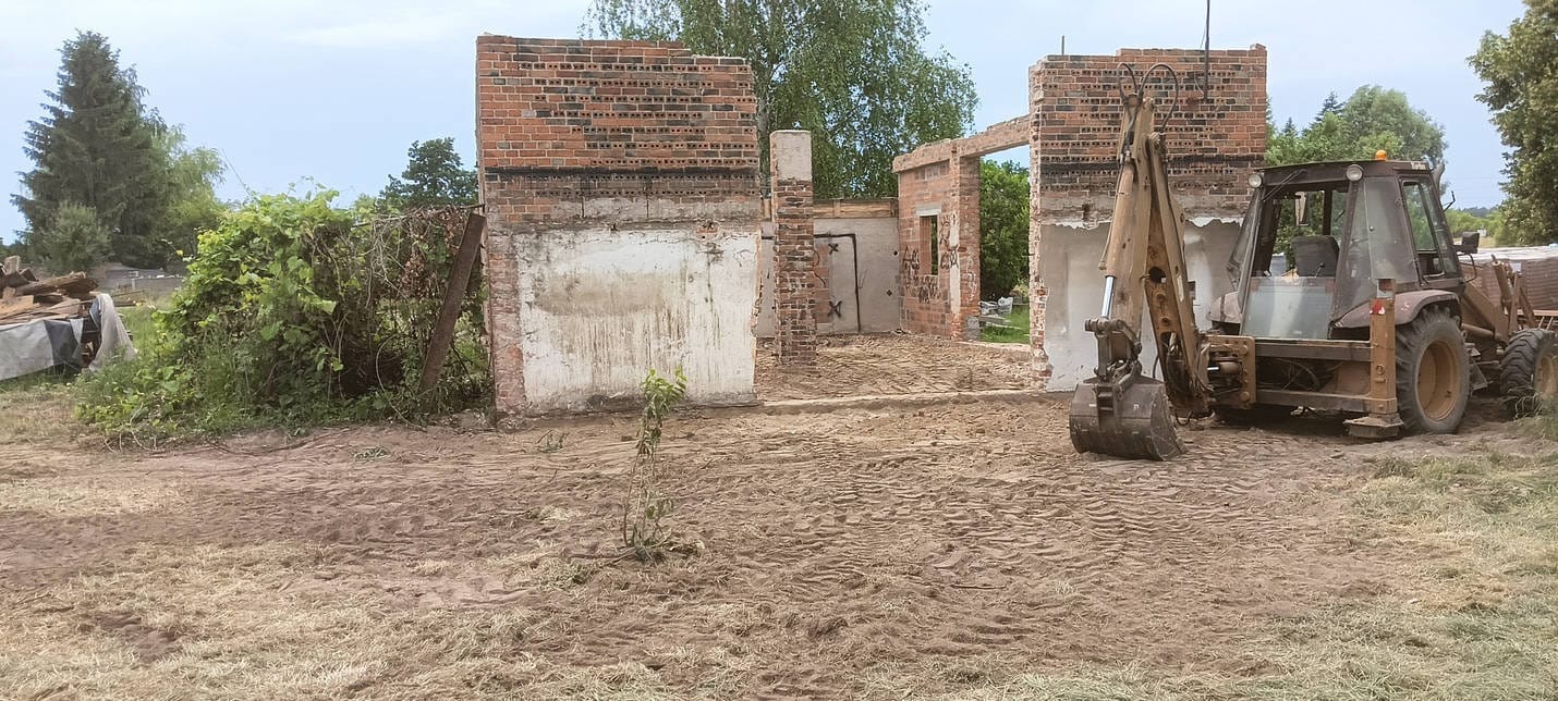Częściowo rozebrany budynek z czerwonej cegły, z widocznym traktorem z przodu, na zewnątrz, w słoneczny dzień. Widoczne ślady pracy ziemnej.