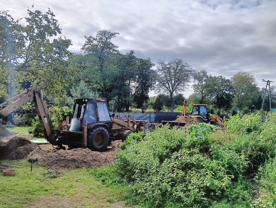 Dwie koparko-ładowarki pracują na zalesionym terenie. Jedna z maszyn nabiera ziemię, druga w tle. Widoczne drzewa i krzewy. Pochmurne niebo.