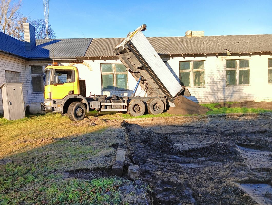 Żółta wywrotka z podniesioną skrzynią wysypuje ziemię na przygotowany teren obok budynku z białej cegły w słoneczny dzień. Widoczne ślady opon i świeżo rozplantowana ziemia.