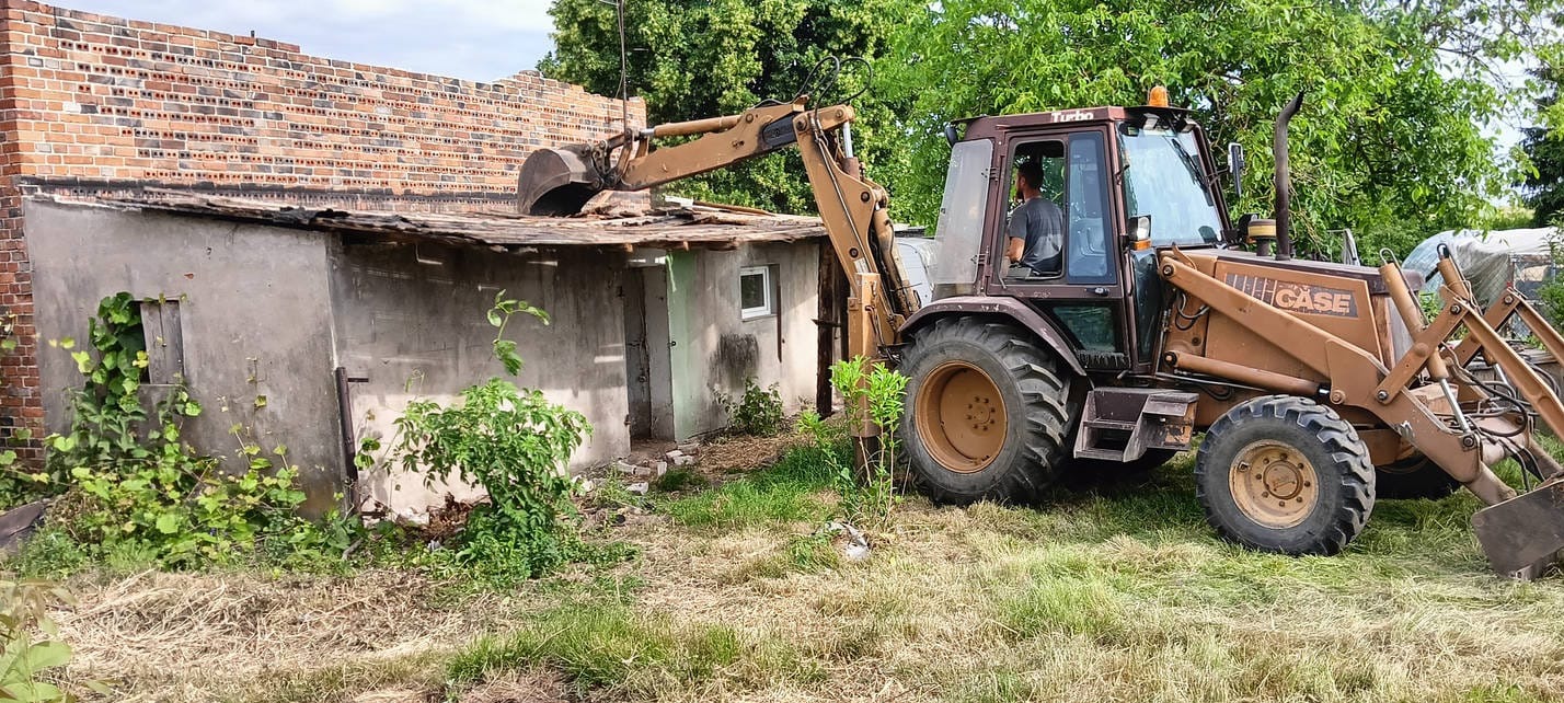 Żółto-brązowa koparko-ładowarka CASE podczas rozbiórki małego, murowanego budynku z ceglanym fragmentem ściany. Operator w kabinie. Widoczne elementy roślinności.