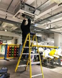 Montaż sufitowej klimatyzacji kasetonowej w biurze. Instalator na drabinie, widoczne elementy instalacji i oświetlenie. Jasne wnętrze z elementami wyposażenia.