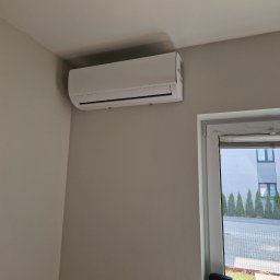 ASPEN Adrian Kosiela - Biały klimatyzator ścienny zamontowany w jasnym pomieszczeniu, widok z dołu, okno z widokiem na zewnątrz po prawej stronie.