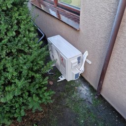 ASPEN Adrian Kosiela - Biały klimatyzator Rotenso zamontowany na zewnątrz budynku, obok zielonego krzewu iglastego i brązowej rury spustowej. Widoczne przewody i elementy montażowe.