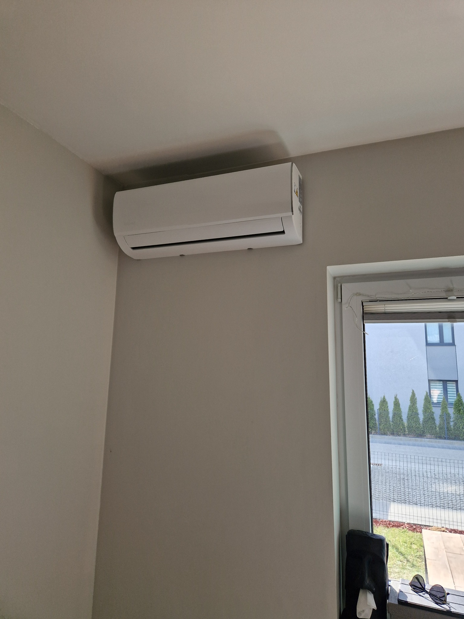 Biały klimatyzator ścienny zamontowany w jasnym pomieszczeniu, widok z dołu, okno z widokiem na zewnątrz po prawej stronie.