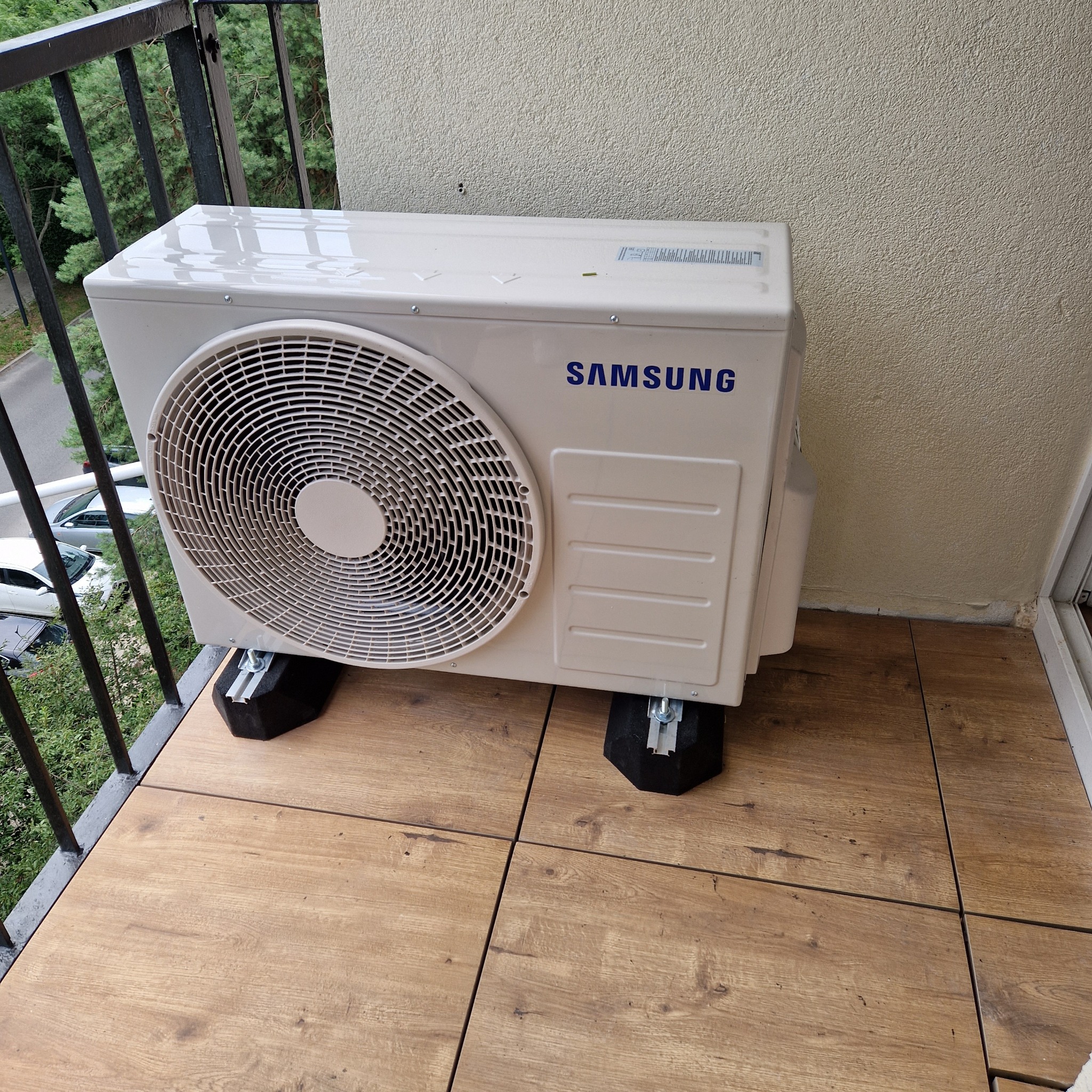 Kompaktowa jednostka zewnętrzna klimatyzacji Samsung na balkonie, zamontowana na czarnych podkładkach antywibracyjnych. Widok z góry, balustrada i otoczenie budynku w tle.