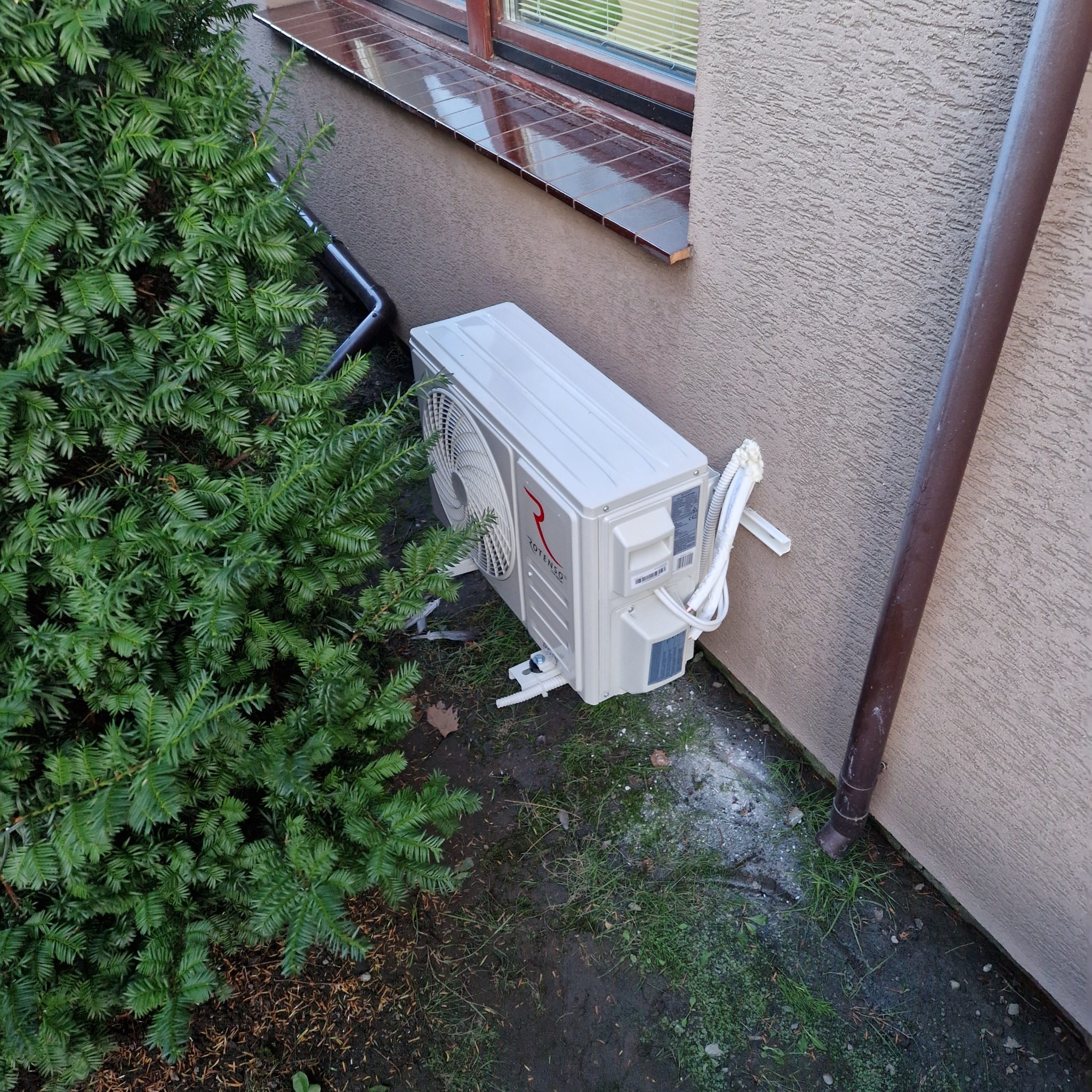 Biały klimatyzator Rotenso zamontowany na zewnątrz budynku, obok zielonego krzewu iglastego i brązowej rury spustowej. Widoczne przewody i elementy montażowe.
