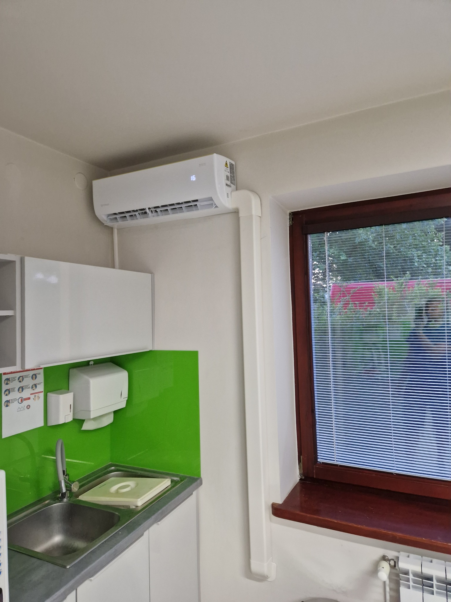 Biały klimatyzator ścienny zamontowany w pomieszczeniu z zielonym panelem, widoczny fragment okna z żaluzjami i grzejnik. Instalacja połączona estetycznymi rurami.