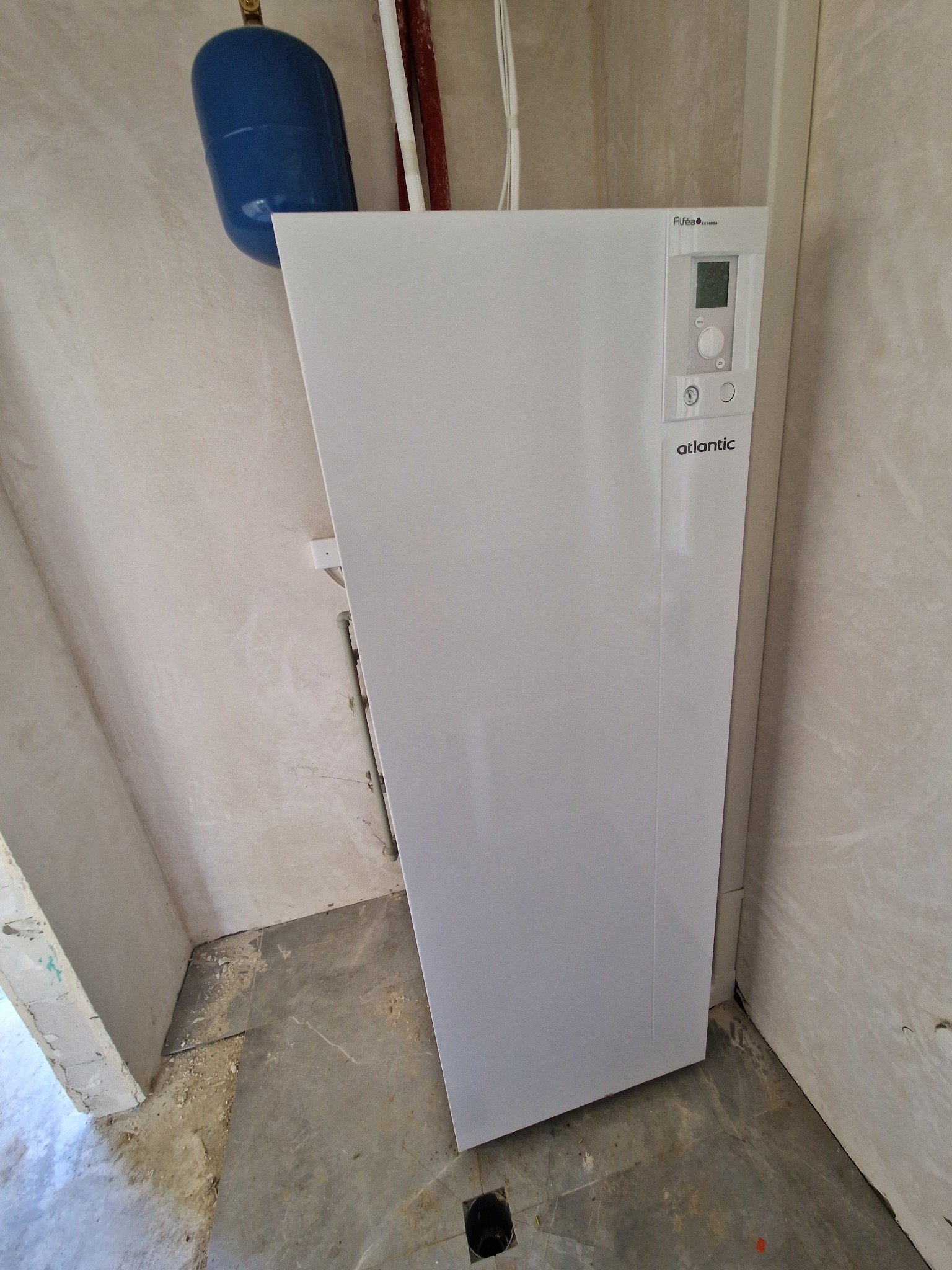 Biała pompa ciepła Atlantic Alfea Extensa, zamontowana w rogu pomieszczenia w trakcie remontu, z widocznym niebieskim zbiornikiem wyrównawczym.