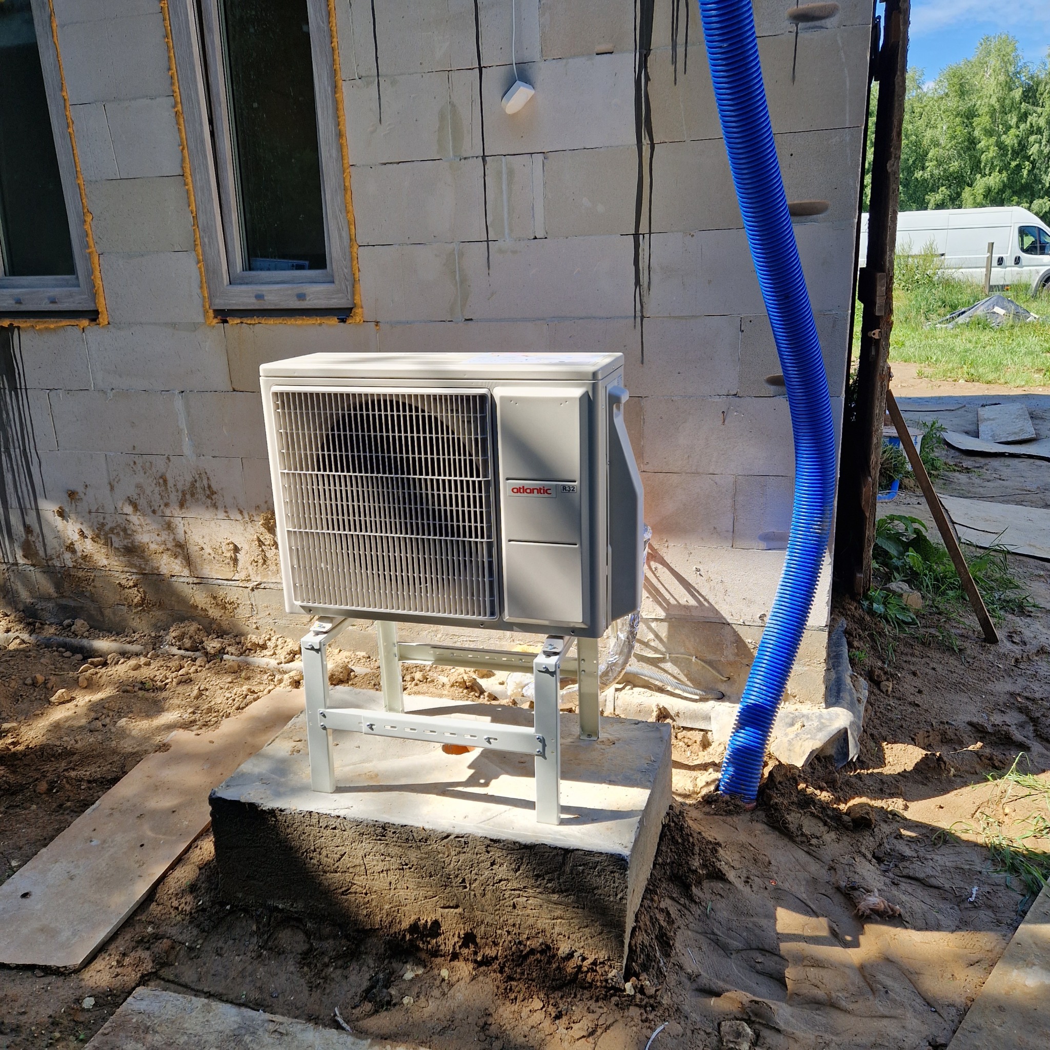 Zewnętrzna jednostka klimatyzacji Atlantic R32 na betonowym fundamencie przy ścianie budynku z pustaków, z widocznymi oknami i niebieskim przewodem wentylacyjnym.