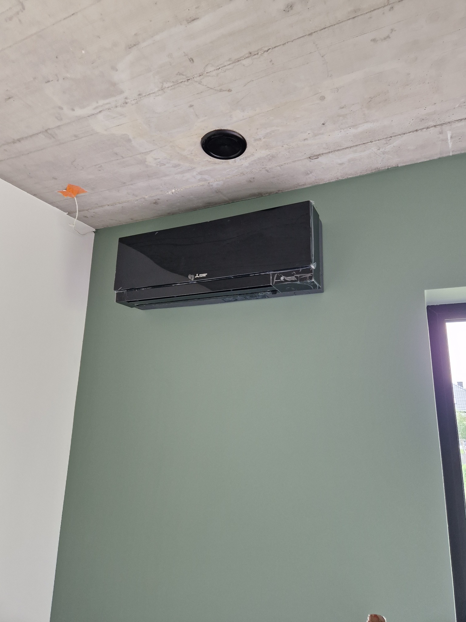Czarny klimatyzator Mitsubishi zamontowany na zielonej ścianie w surowym wnętrzu. Widoczny betonowy sufit z otworem na oświetlenie i wystające kable.