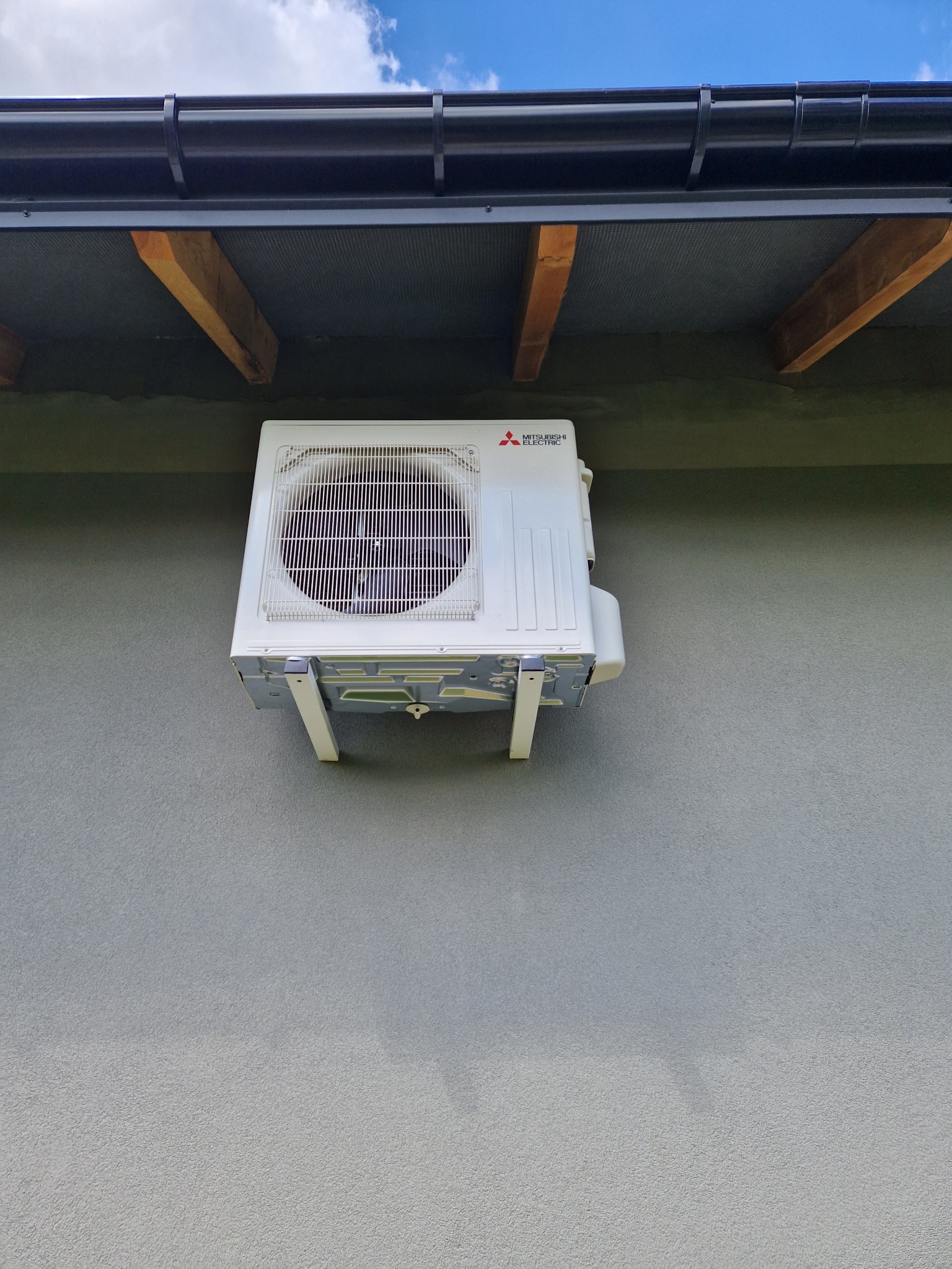 Biała jednostka zewnętrzna klimatyzacji Mitsubishi Electric zamontowana na szarej ścianie budynku, widoczna z dołu, z fragmentem dachu i niebem w tle.