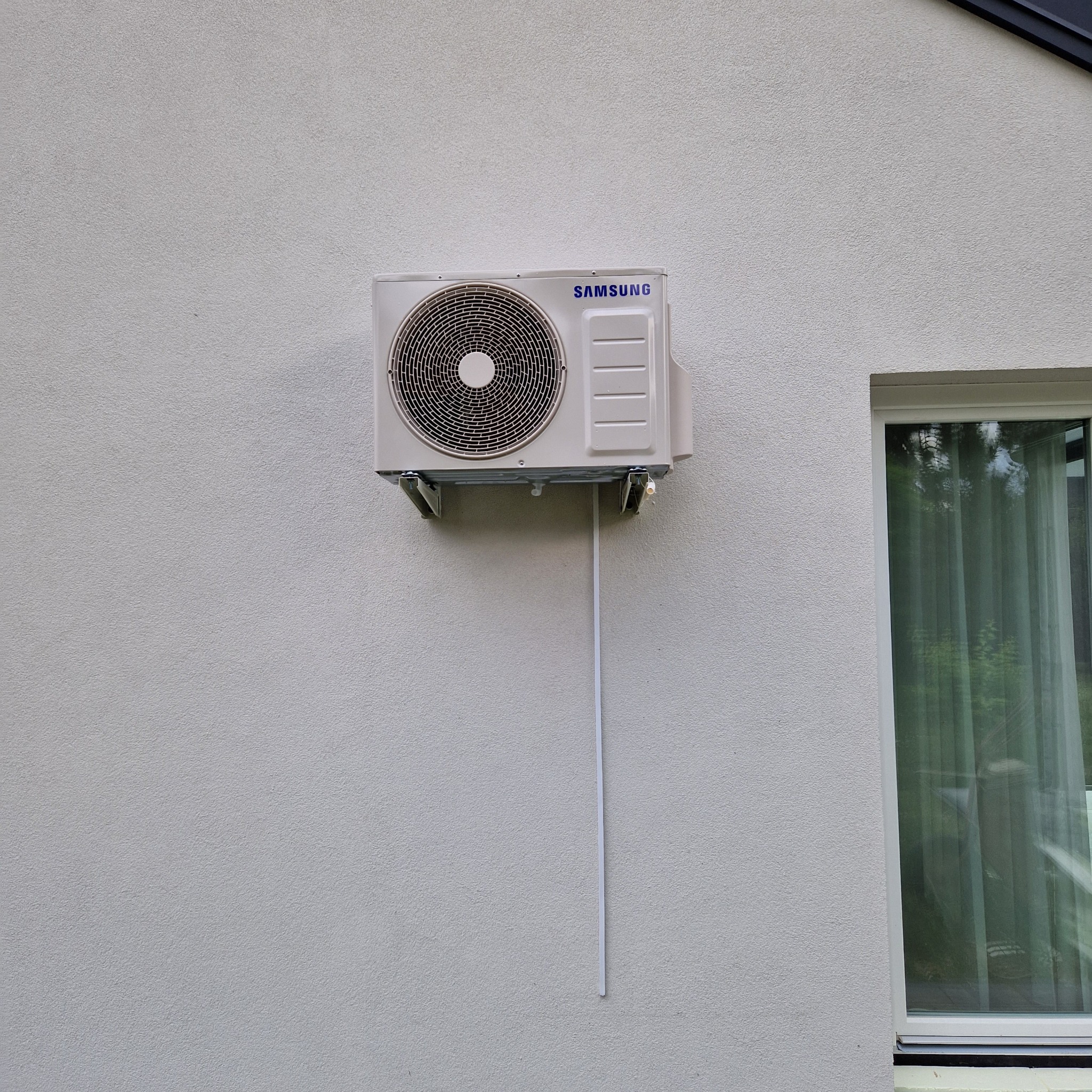 Zamontowana jednostka zewnętrzna klimatyzacji Samsung na jasnej, teksturowanej ścianie budynku, obok okna z zielonymi zasłonami. Widoczne metalowe wsporniki i biała rurka odprowadzająca.