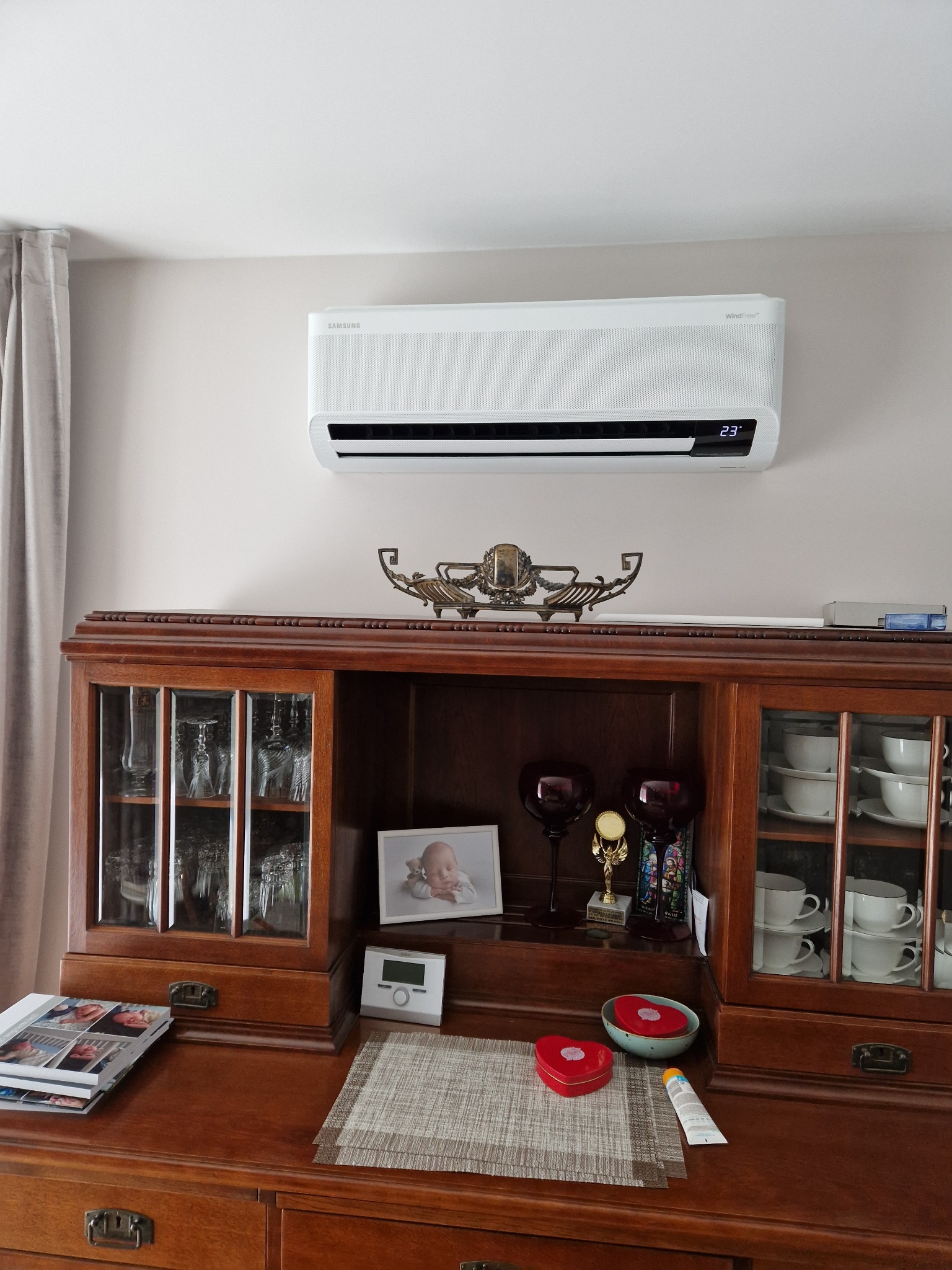 Biały klimatyzator Samsung WindFree zamontowany nad drewnianym kredensem w salonie. Na kredensie dekoracje, zdjęcia i termostat. Widoczny fragment zasłony.