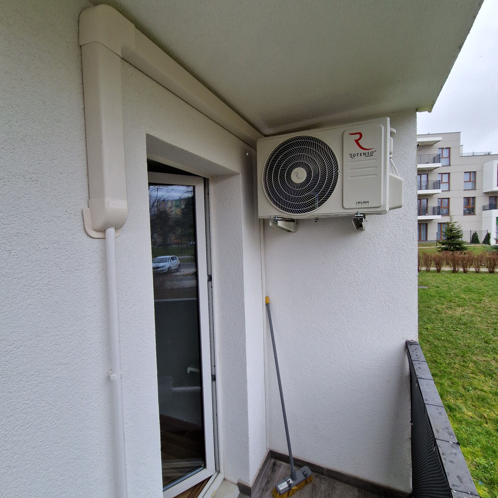 Biała jednostka zewnętrzna klimatyzacji Rotenso zamontowana na balkonie bloku mieszkalnego, widoczne rury odprowadzające. Na podłodze stoi miotła.