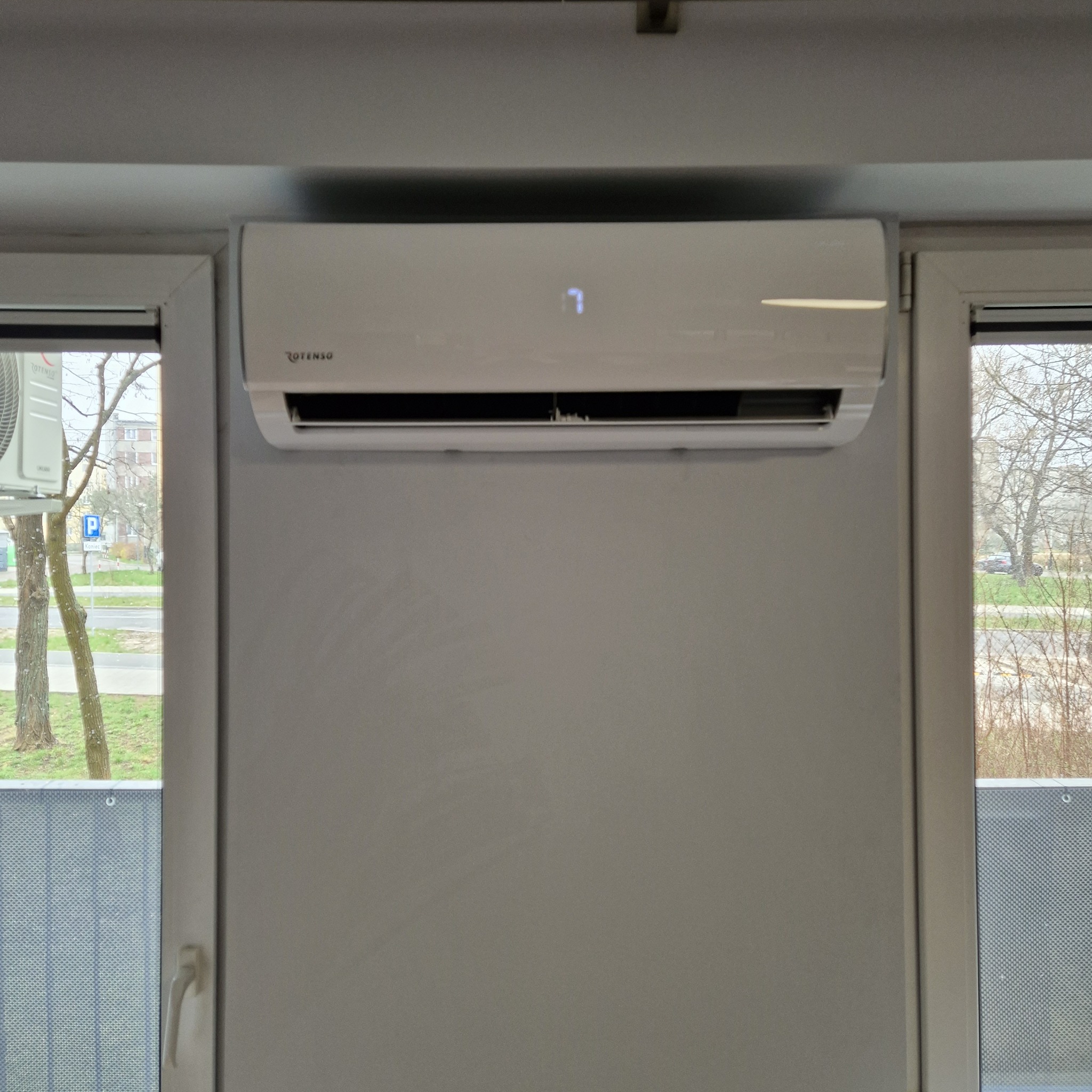 Biały klimatyzator Rotenso zamontowany nad oknem, widoczny wyświetlacz z cyfrą '7'. Za oknem drzewa i budynki w tle. Minimalistyczny design.