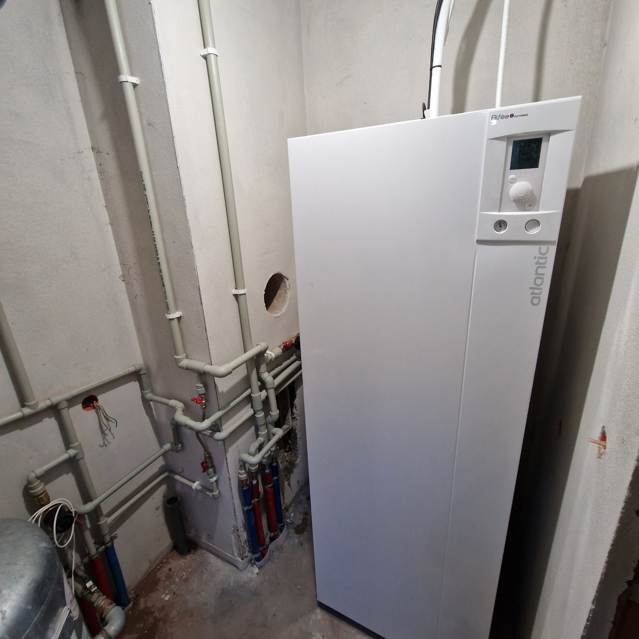 Biała pompa ciepła Atlantic z podłączonymi rurami w surowym pomieszczeniu. Widoczne elementy instalacji i otwór w ścianie.