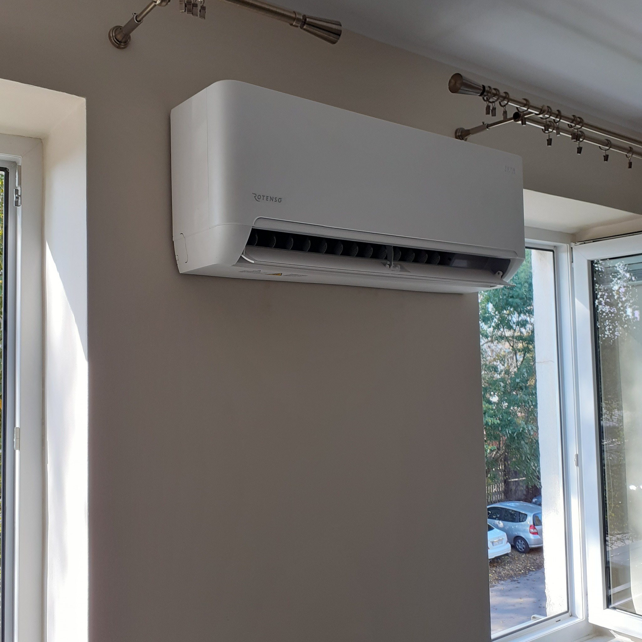 Biały klimatyzator Rotenso Delta zamontowany na ścianie w pokoju, widok z boku, okno z widokiem na zewnątrz po prawej stronie.