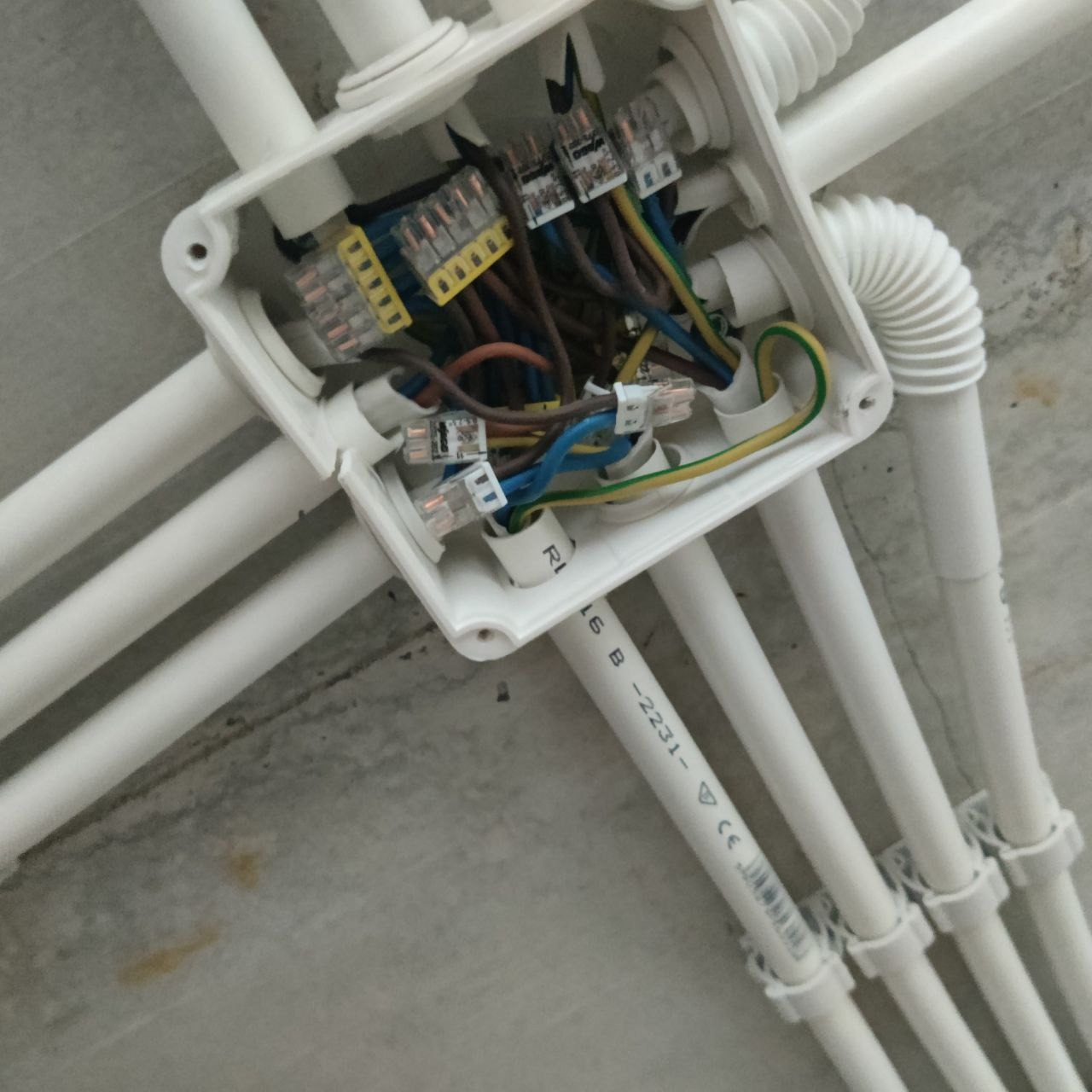 Biała puszka elektryczna z widocznymi przewodami i złączkami WAGO na szarym suficie, otoczona białymi rurkami instalacyjnymi.