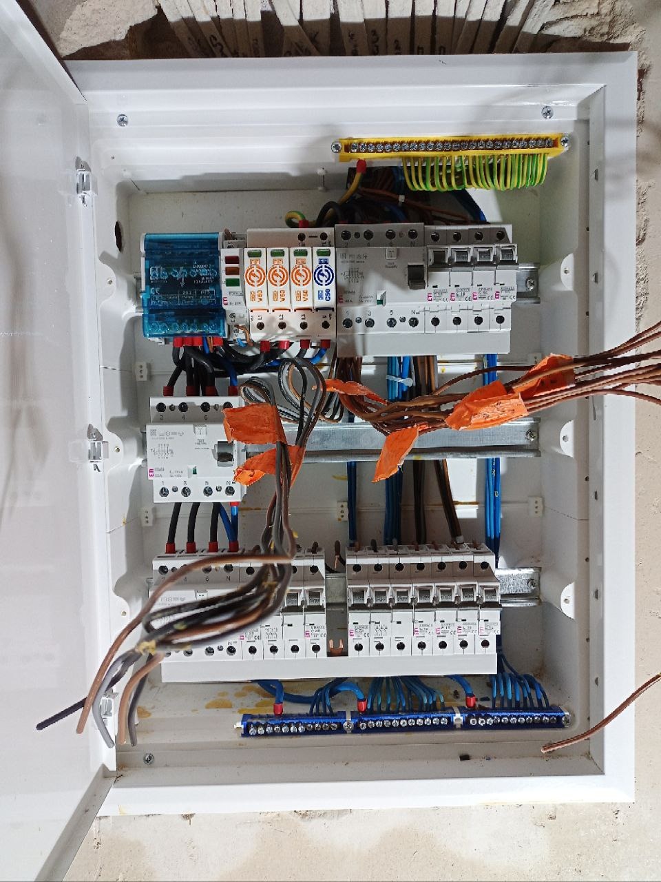 Otwarta skrzynka elektryczna z zabezpieczeniami, przewodami i listwami zaciskowymi, widok z góry, przewody zabezpieczone pomarańczową taśmą.