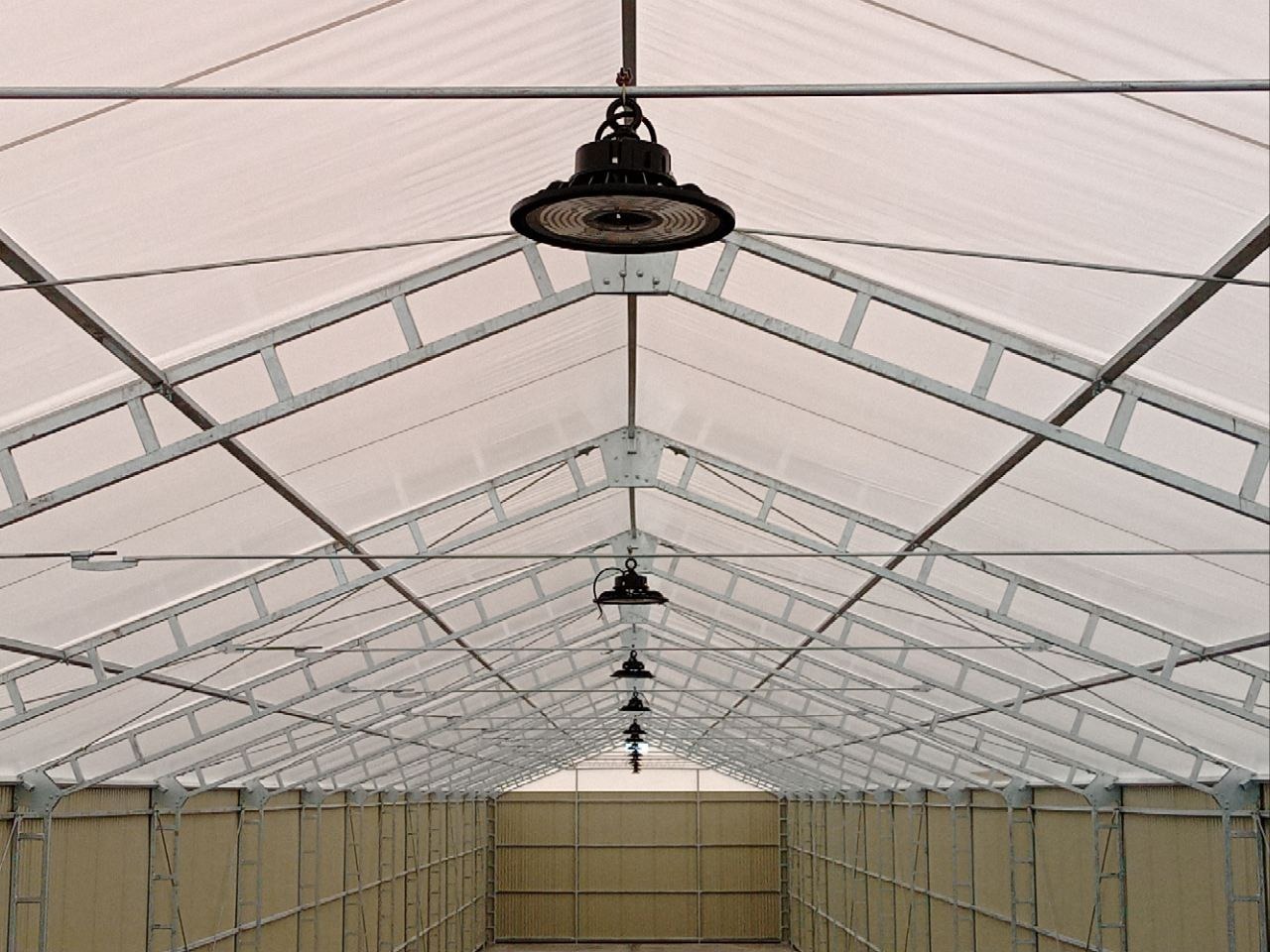 Wnętrze hali z metalową konstrukcją dachu i rzędem okrągłych lamp LED, oświetlających przestrzeń. Widok perspektywiczny w głąb hali.