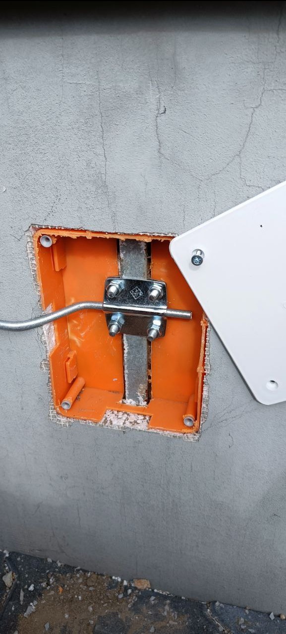 Pomarańczowa puszka instalacyjna w szarej ścianie z zamontowanym metalowym uchwytem i wystającym przewodem, częściowo zakryta białą obudową. Widoczne ślady po montażu.