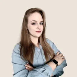 Portret kobiety z brązowymi włosami, ubranej w błękitną koszulę, z założonymi rękami i zegarkiem na nadgarstku, na beżowym tle.