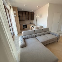 Apartamenty V-Kiff - Jasne wnętrze z narożnikiem w odcieniach szarości, parkietem w jodełkę i kuchnią w tle. Widok z okna z delikatną firanką. Minimalistyczny design.