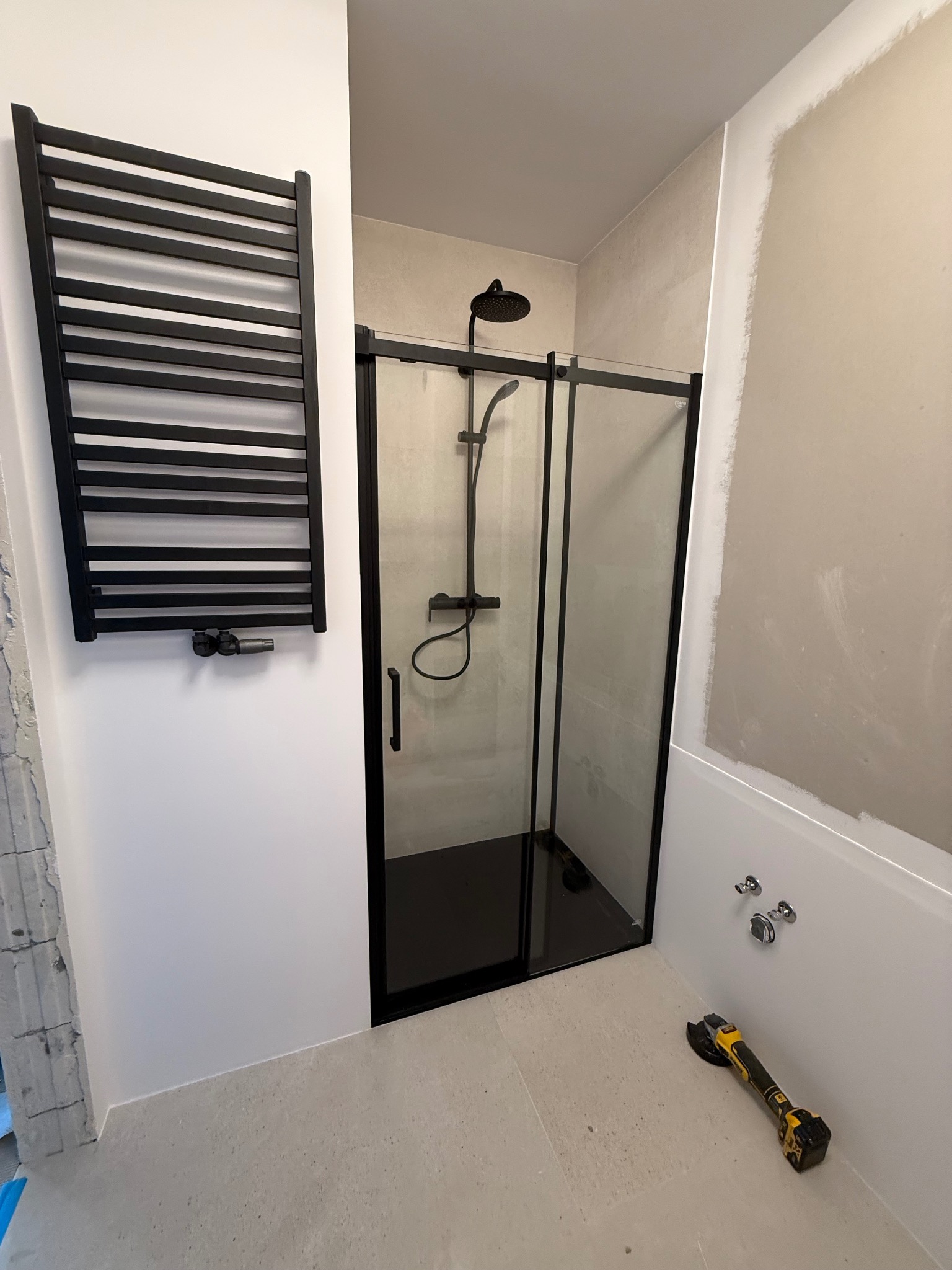 Nowoczesna łazienka po remoncie: czarna kabina prysznicowa, grzejnik drabinkowy i płytki w odcieniach szarości. Na podłodze leży szlifierka kątowa.