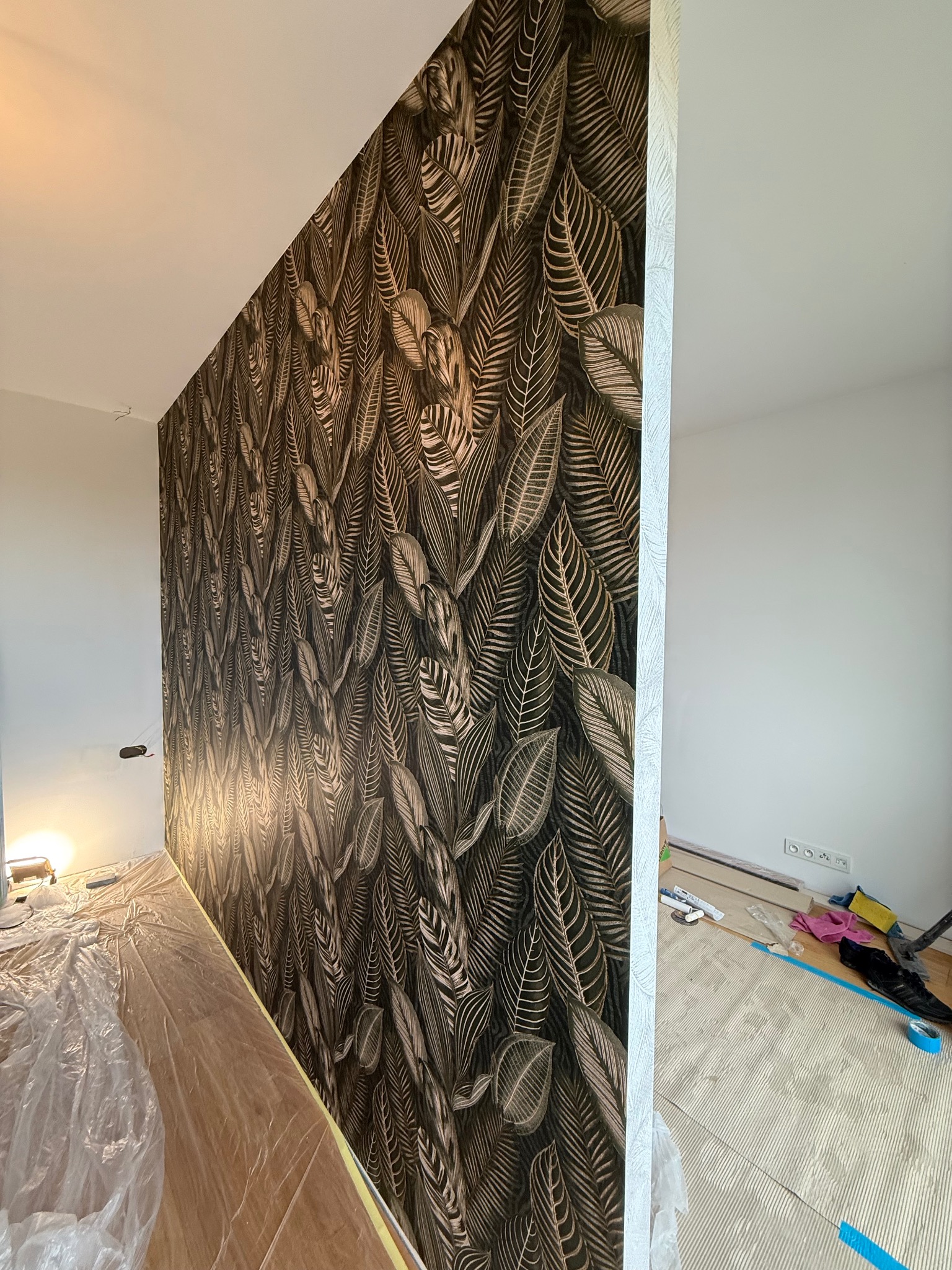 Ściana z tapetą w liście, widok z boku, z zabezpieczoną podłogą. Fragment białej ściany po prawej. Dekoracja wnętrza w trakcie prac wykończeniowych.