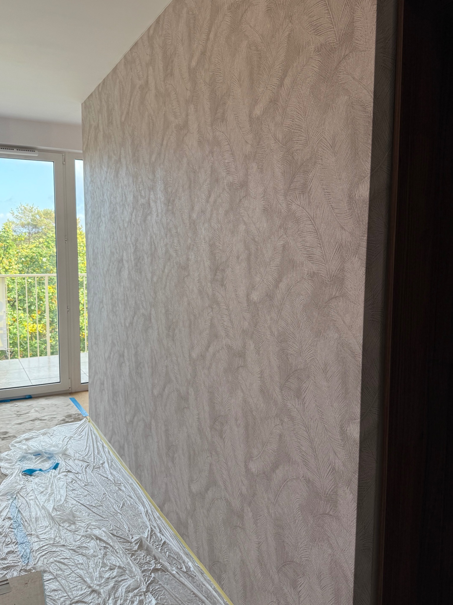 Ściana z tapetą w delikatny wzór piór, zabezpieczona folią malarską. Widoczne okno balkonowe i fragment drzwi. Jasne, naturalne światło.