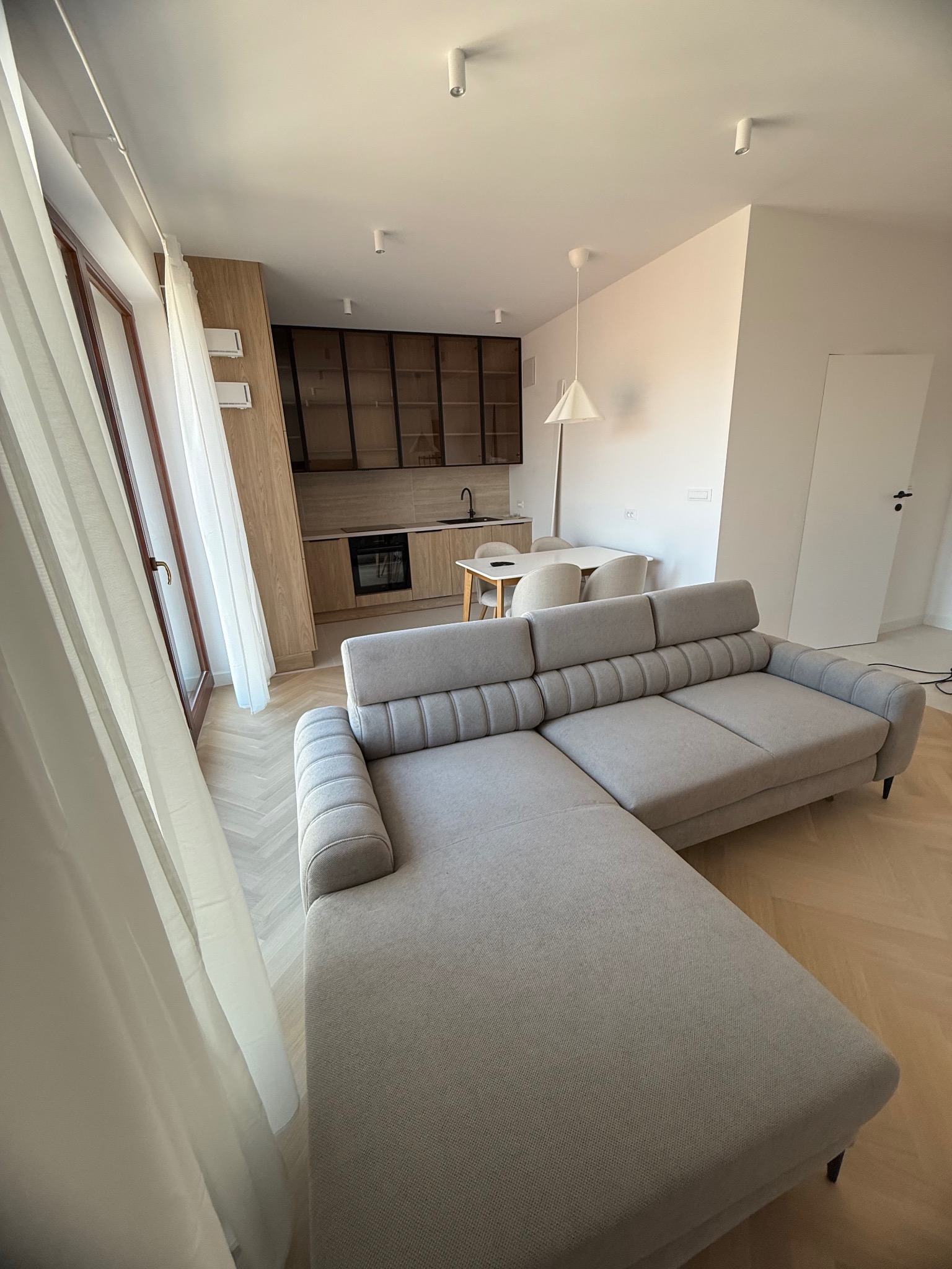 Jasne wnętrze z narożnikiem w odcieniach szarości, parkietem w jodełkę i kuchnią w tle. Widok z okna z delikatną firanką. Minimalistyczny design.