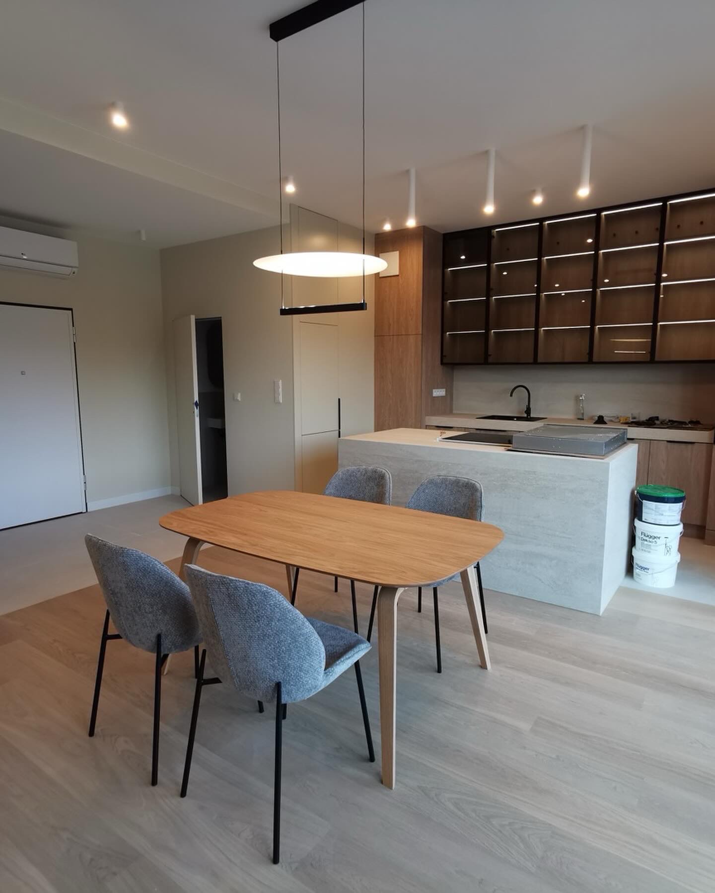 Nowoczesna kuchnia z wyspą i drewnianym stołem z szarymi krzesłami. Widoczne farby w tle. Oświetlenie sufitowe punktowe i wiszące. Jasna podłoga.