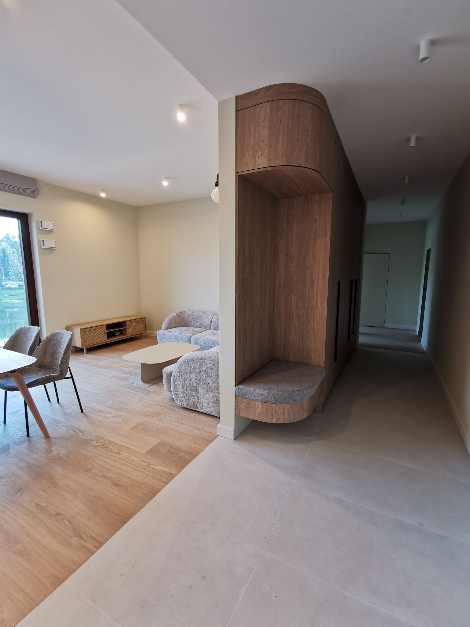 Nowoczesne wnętrze: salon z drewnianą zabudową, szarą sofą i stołem. Widoczny korytarz z jasnymi płytkami. Minimalistyczny design, ciepłe oświetlenie.