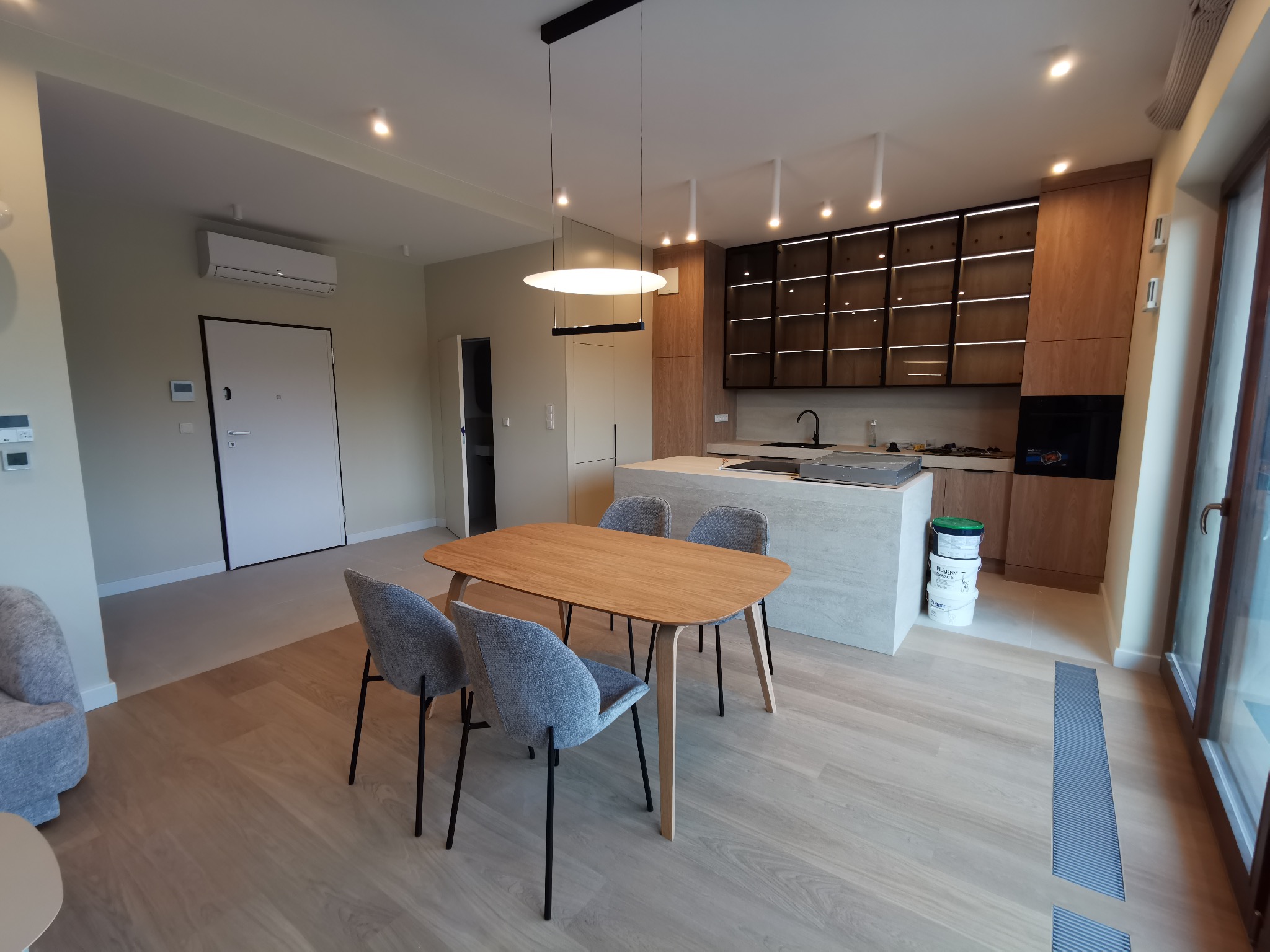 Nowoczesna kuchnia z wyspą i drewnianymi szafkami podświetlonymi LED, stół z szarymi krzesłami na parkiecie. Widoczne farby w wiadrach, klimatyzator i drzwi.