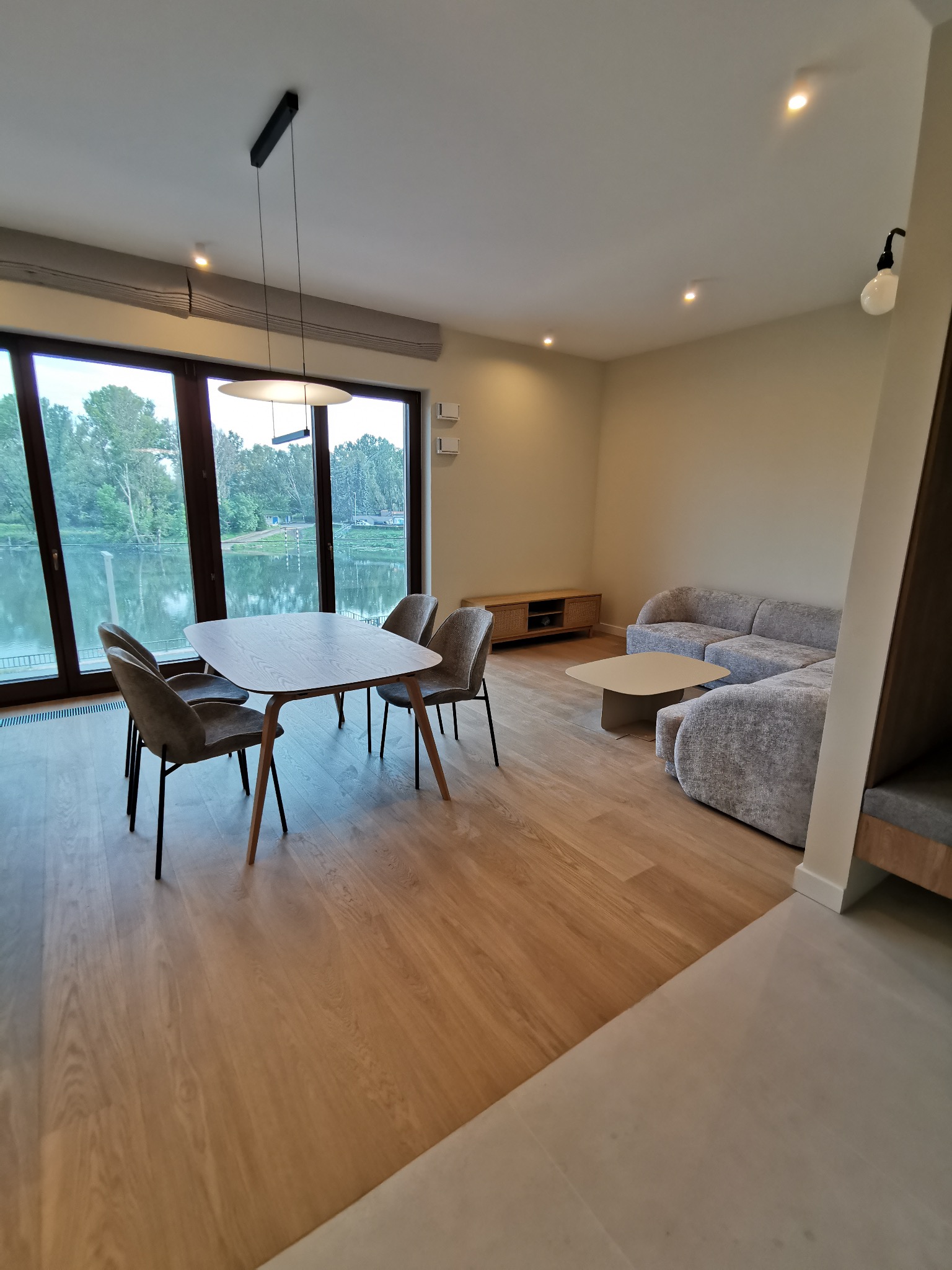 Jasne wnętrze salonu z dużym oknem na jezioro. Stół z krzesłami, sofa i drewniana podłoga tworzą nowoczesną przestrzeń. Minimalistyczne oświetlenie.