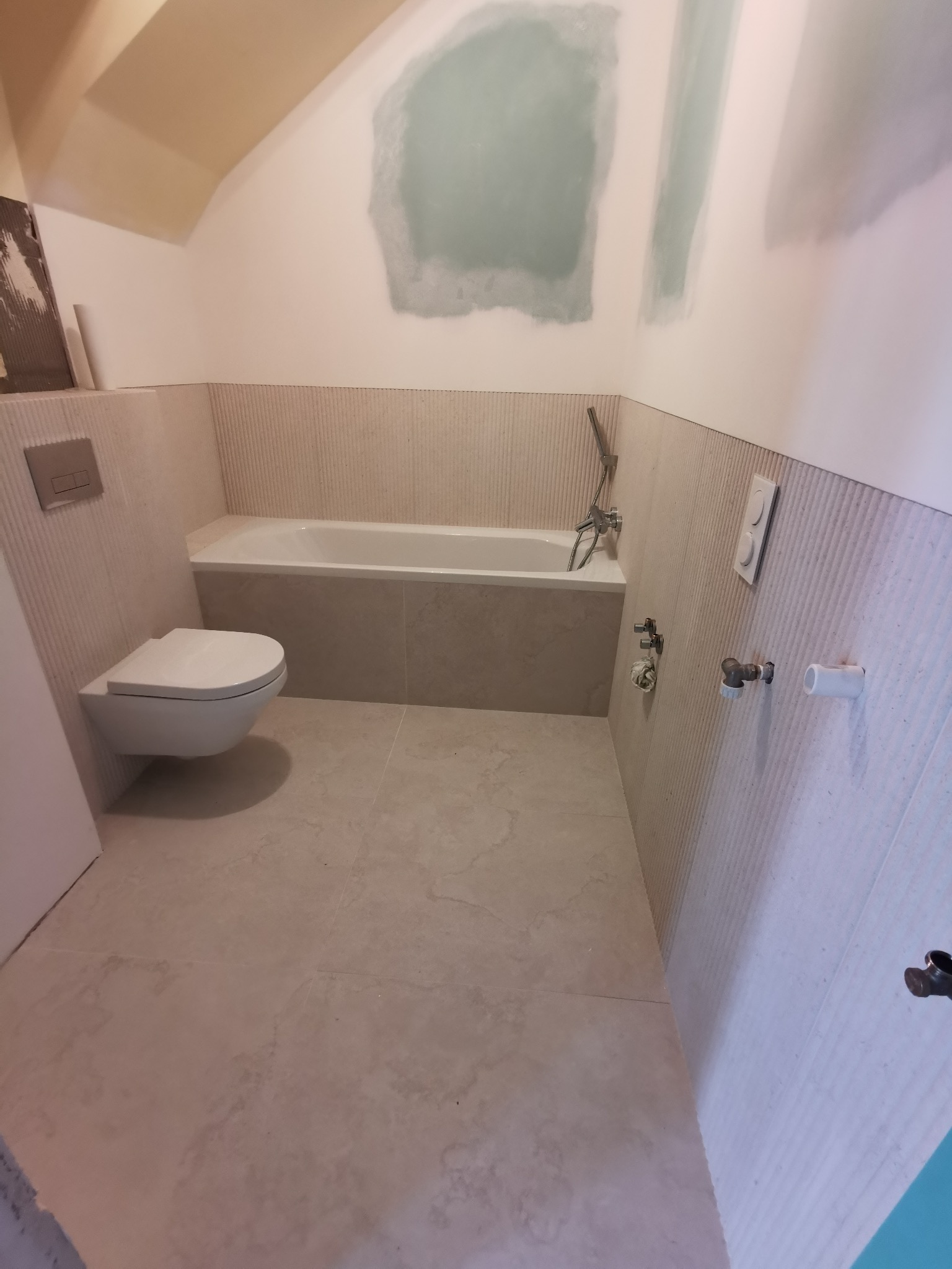 Jasna łazienka w trakcie wykańczania: beżowe płytki ścienne z subtelnym wzorem, podwieszana toaleta, wanna i fragment ściany z widocznymi śladami gładzi.