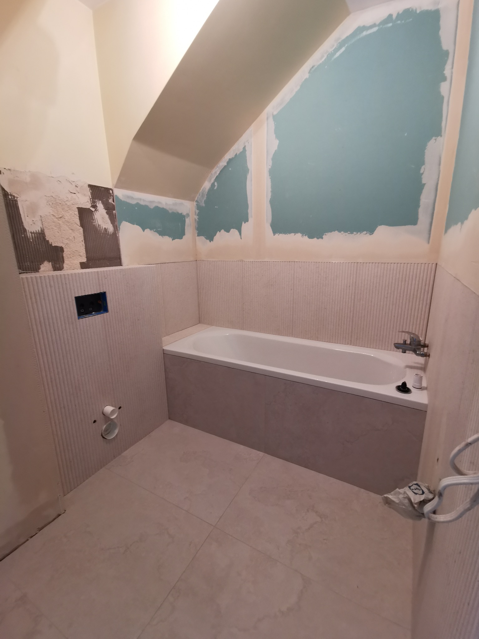 Remont łazienki w trakcie: częściowo wyłożona płytkami ściana z wanną, fragmenty ścian z płytami karton-gips, widoczne elementy instalacji. Ukończona podłoga z płytek.