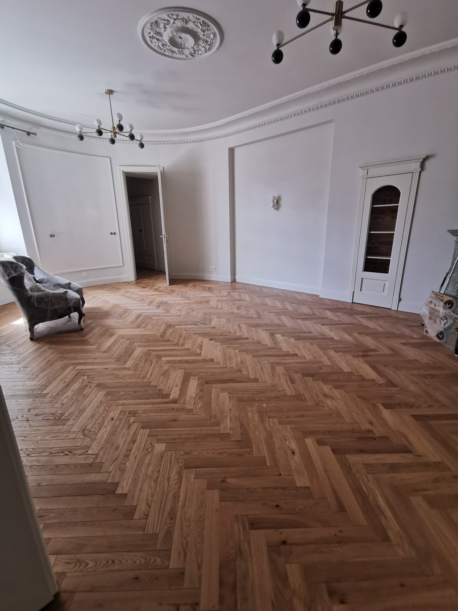 Parkiet ułożony w jodełkę francuską w przestronnym, jasnym salonie z dekoracyjną sztukaterią i nowoczesnym oświetleniem. Widoczny fotel i drzwi.