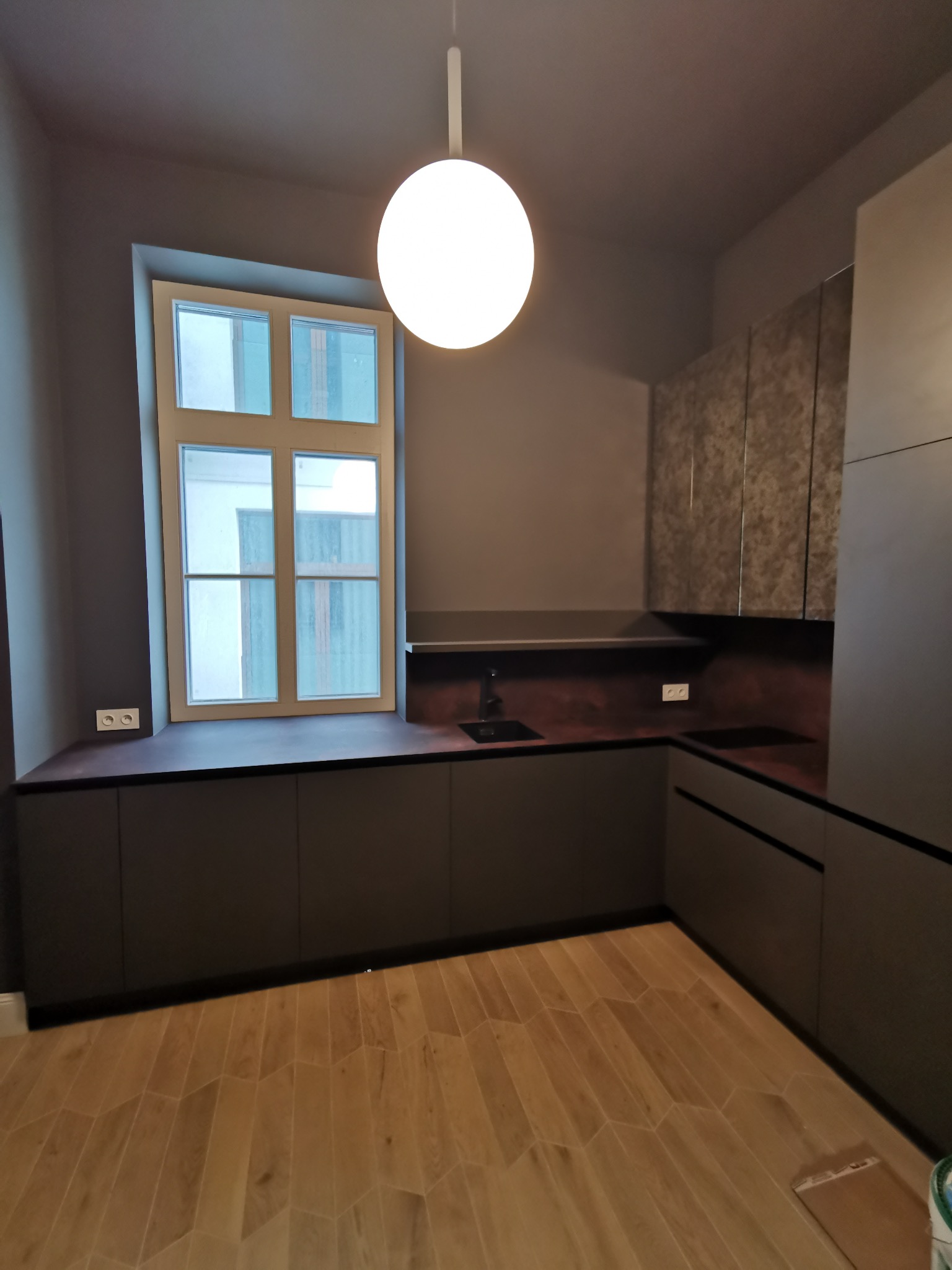 Nowoczesna kuchnia w odcieniach szarości z drewnianą podłogą w jodełkę. Widoczne okno z białymi ramami i minimalistyczna lampa sufitowa.