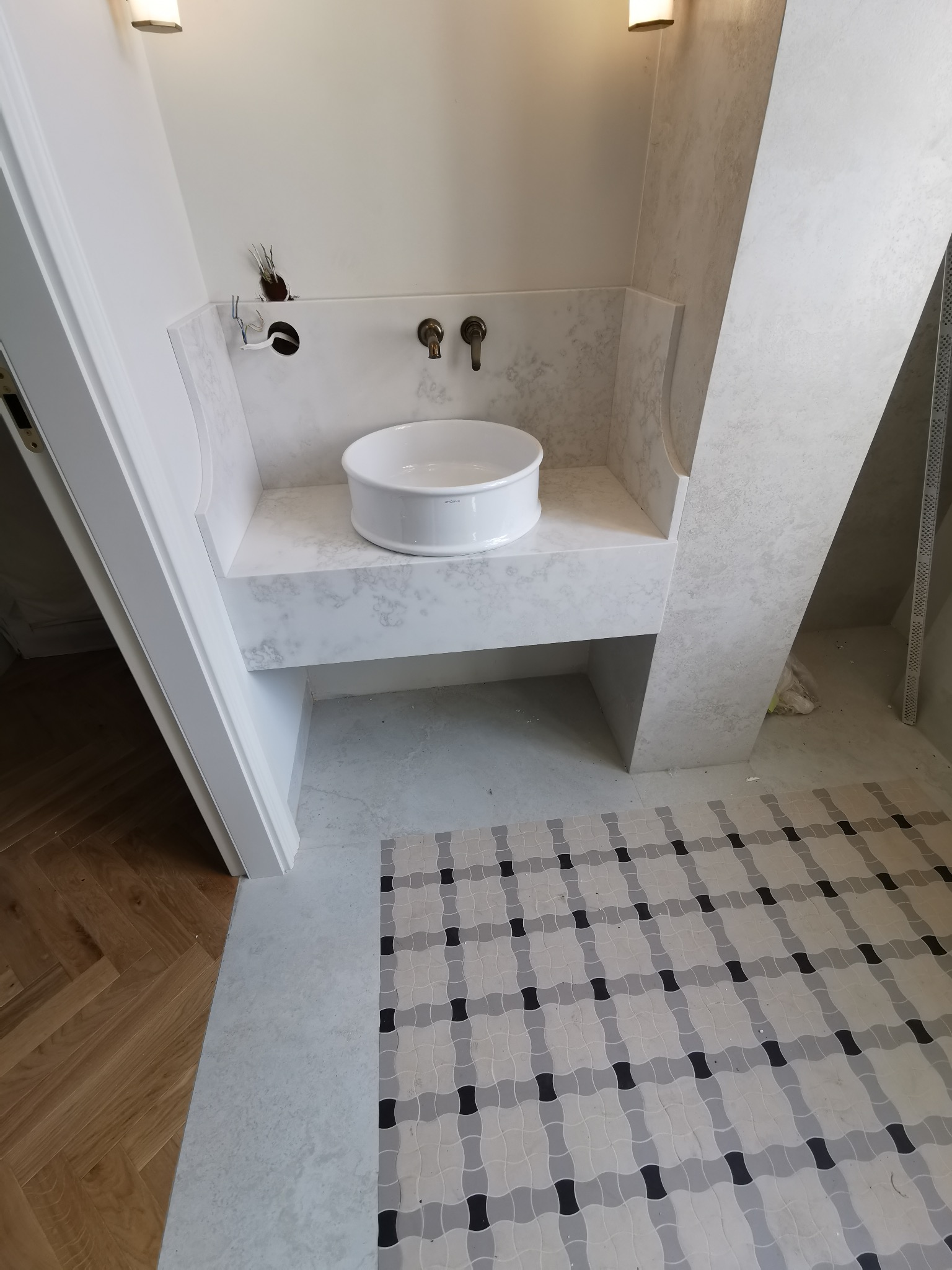 Nowoczesna łazienka w trakcie wykończenia: biała umywalka nablatowa, marmurowy blat, mozaika podłogowa w odcieniach szarości i czerni, widoczne elementy instalacji.