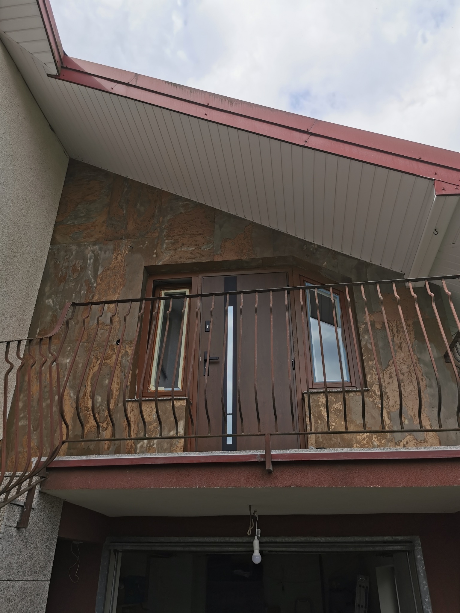 Balkon z brązową balustradą i drzwiami balkonowymi na tle ściany wykończonej płytami imitującymi kamień. Widok z dołu, fragment elewacji budynku.