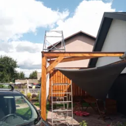 Drewniana pergola z szarą osłoną przeciwsłoneczną w ogrodzie, widoczna drabina i rusztowanie, częściowo zasłonięta przez samoch&oacute;d. Błękitne niebo w tle.