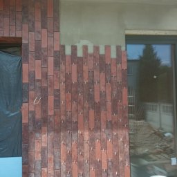 MASTERKOLOS SPÓŁKA Z OGRANICZONĄ ODPOWIEDZIALNOŚCIĄ - Ceglana okładzina elewacyjna w trakcie montażu, widoczne krzyżyki dystansowe. Część ściany nieotynkowana, okno z odbiciem otoczenia, wiadro i narzędzia.