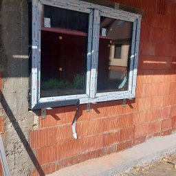 MASTERKOLOS SPÓŁKA Z OGRANICZONĄ ODPOWIEDZIALNOŚCIĄ - Montaż okna PCV w surowym murze z czerwonej cegły. Widoczne uchwyty montażowe i pianka uszczelniająca. Okno dwukomorowe z szybami. Etap prac wykończeniowych.