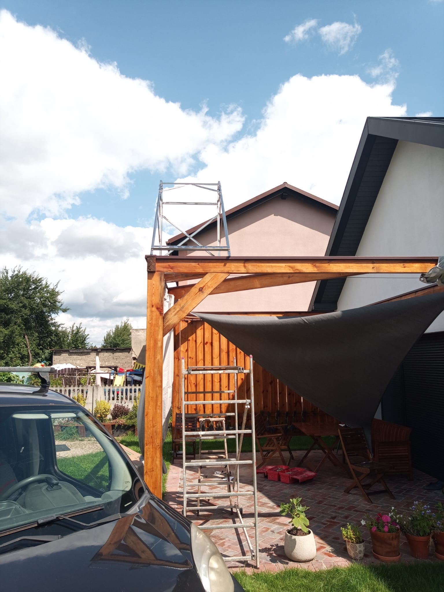 Drewniana pergola z szarą osłoną przeciwsłoneczną w ogrodzie, widoczna drabina i rusztowanie, częściowo zasłonięta przez samochód. Błękitne niebo w tle.