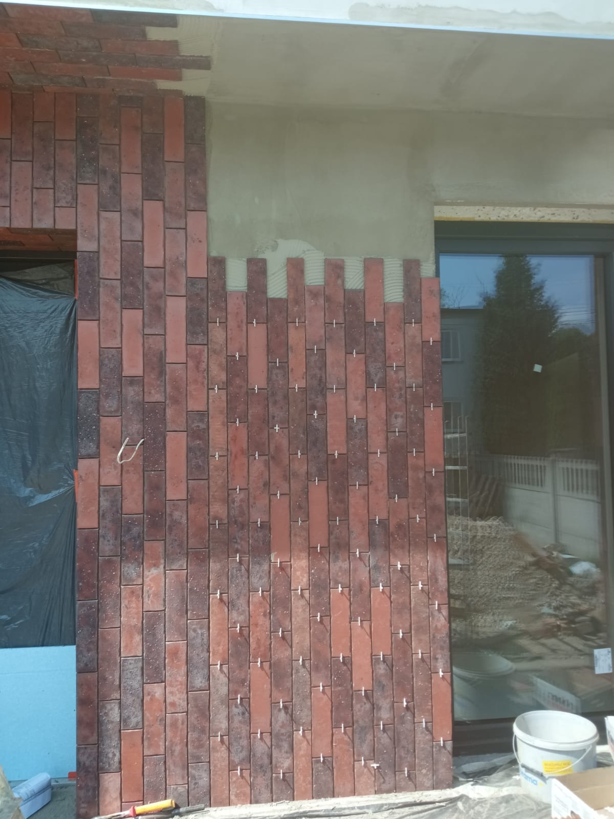 Ceglana okładzina elewacyjna w trakcie montażu, widoczne krzyżyki dystansowe. Część ściany nieotynkowana, okno z odbiciem otoczenia, wiadro i narzędzia.