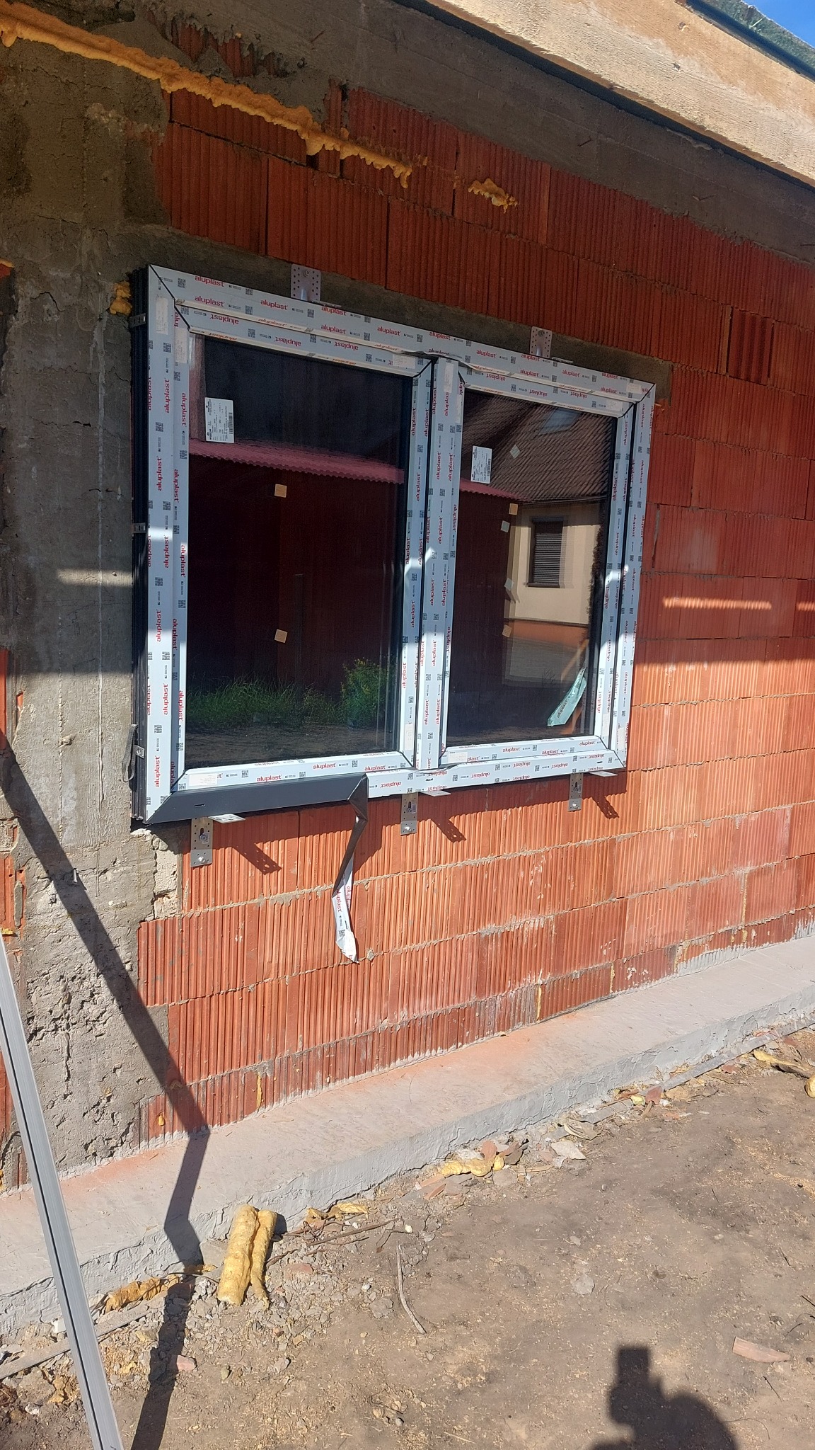 Montaż okna PCV w surowym murze z czerwonej cegły. Widoczne uchwyty montażowe i pianka uszczelniająca. Okno dwukomorowe z szybami. Etap prac wykończeniowych.